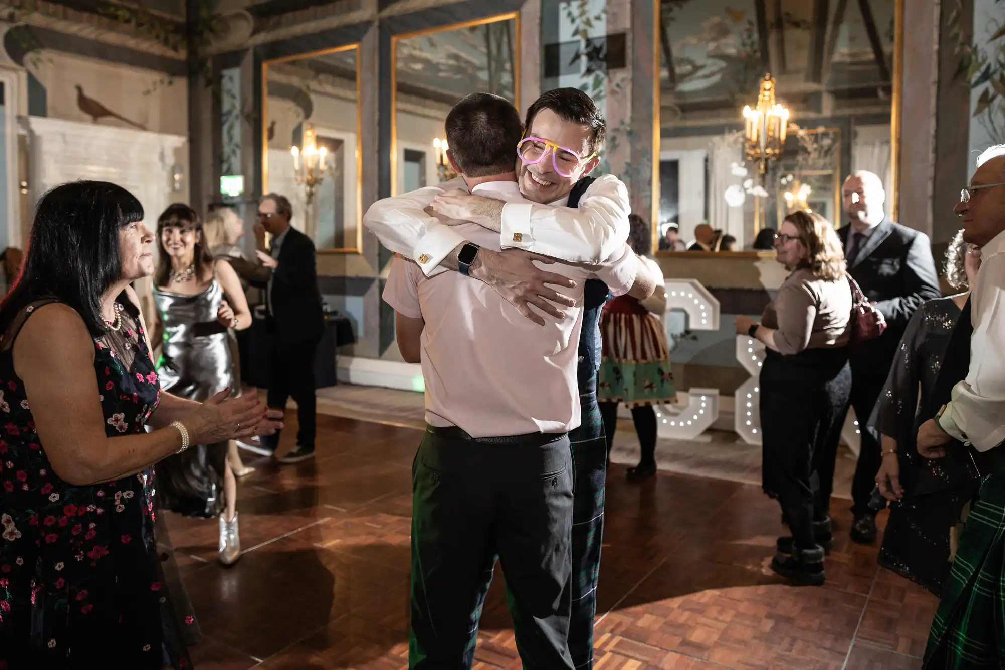 Two people embrace in a hug on a dance floor at a lively indoor gathering. Others are dancing or standing around them. One person wears pink neon glasses. The room has ornate decor and chandeliers.