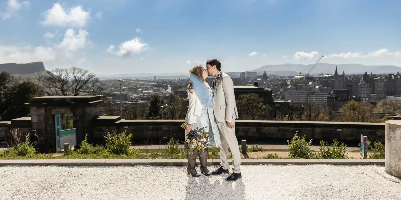 A couple dressed in light-colored formal attire shares a kiss outdoors with a cityscape and clear sky in the background.