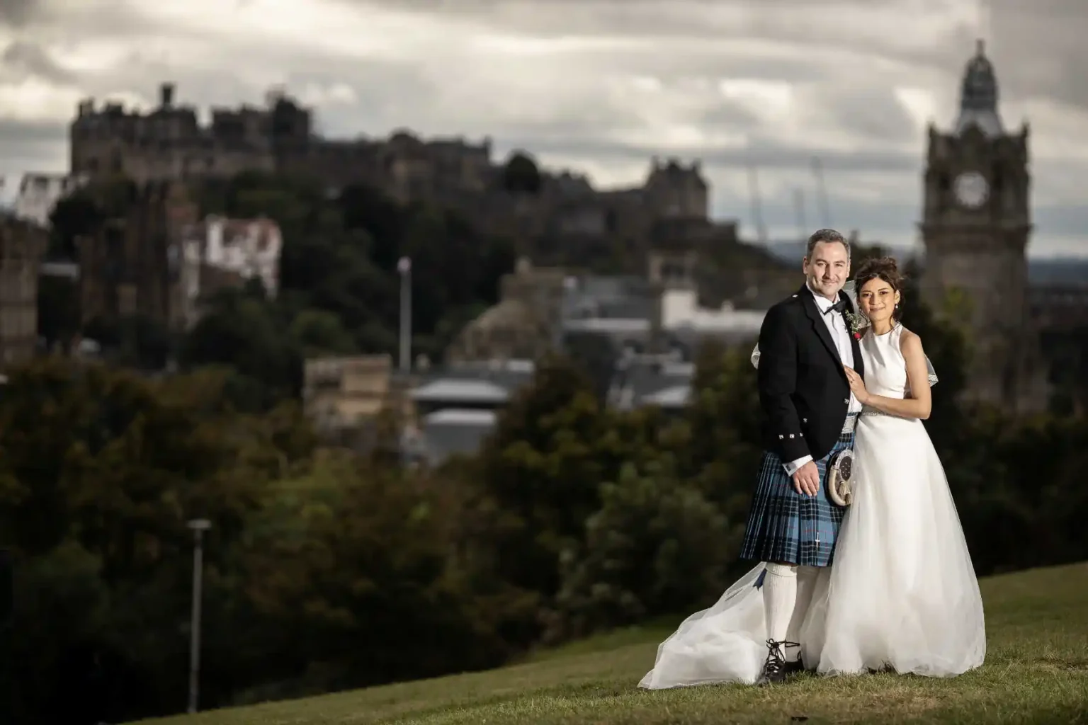 A bride and groom stand together on a grassy hill, with historic stone buildings and a clock tower in the background under a cloudy sky.