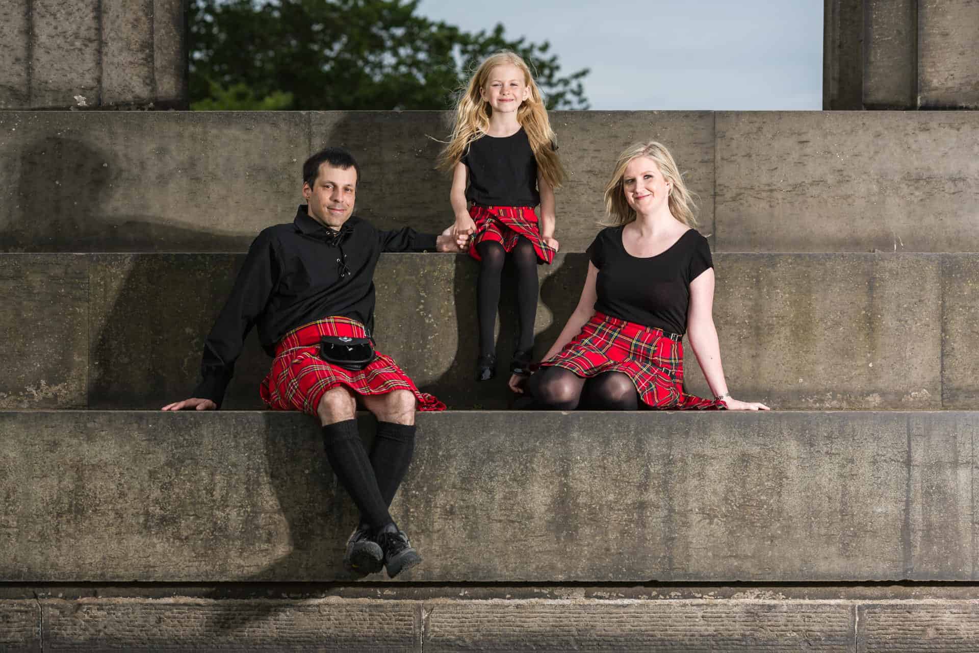 Three people sit on large stone steps, all wearing black tops and red tartan kilts; two adults flank a child in the middle. Trees are visible in the background.