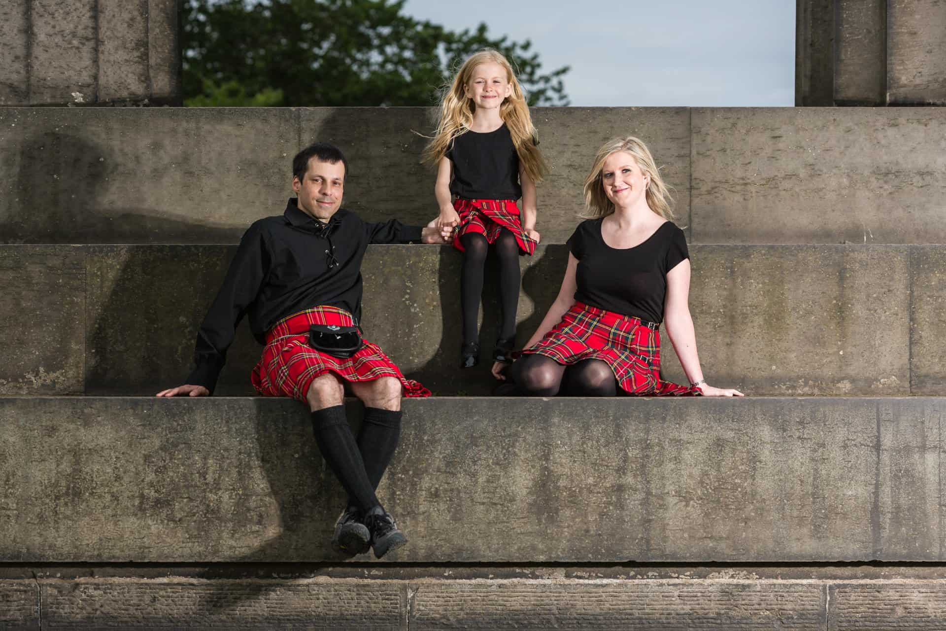Three people sit on large stone steps, all wearing black tops and red tartan kilts; two adults flank a child in the middle. Trees are visible in the background.