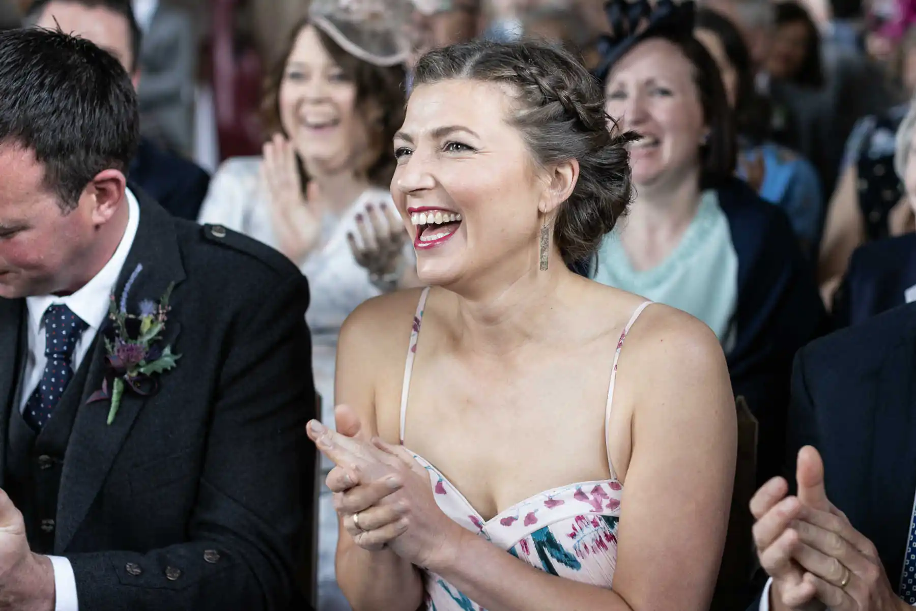 Candid laughter and joy at Rosebery Steading unplugged wedding ceremony: A woman in a floral dress claps and smiles in a crowd, with people around her also clapping and smiling.