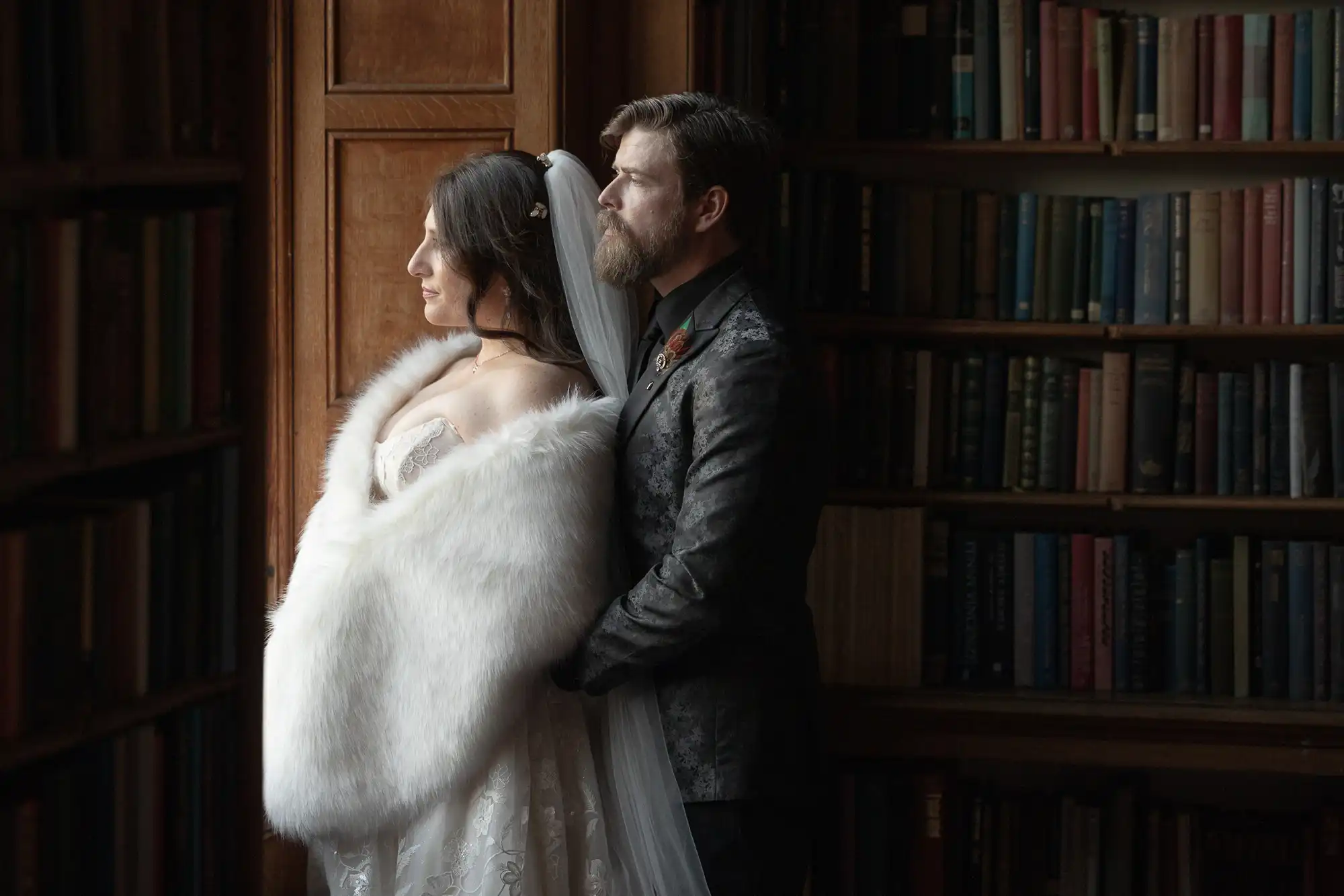 A bride and groom stand together in formal attire beside a bookshelf, gazing out of a window with soft natural light illuminating them.