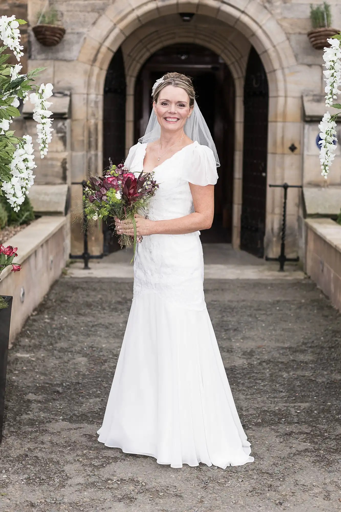 A bride in a white dress and veil holds a bouquet, smiling in front of an ornate stone archway adorned with white flowers.