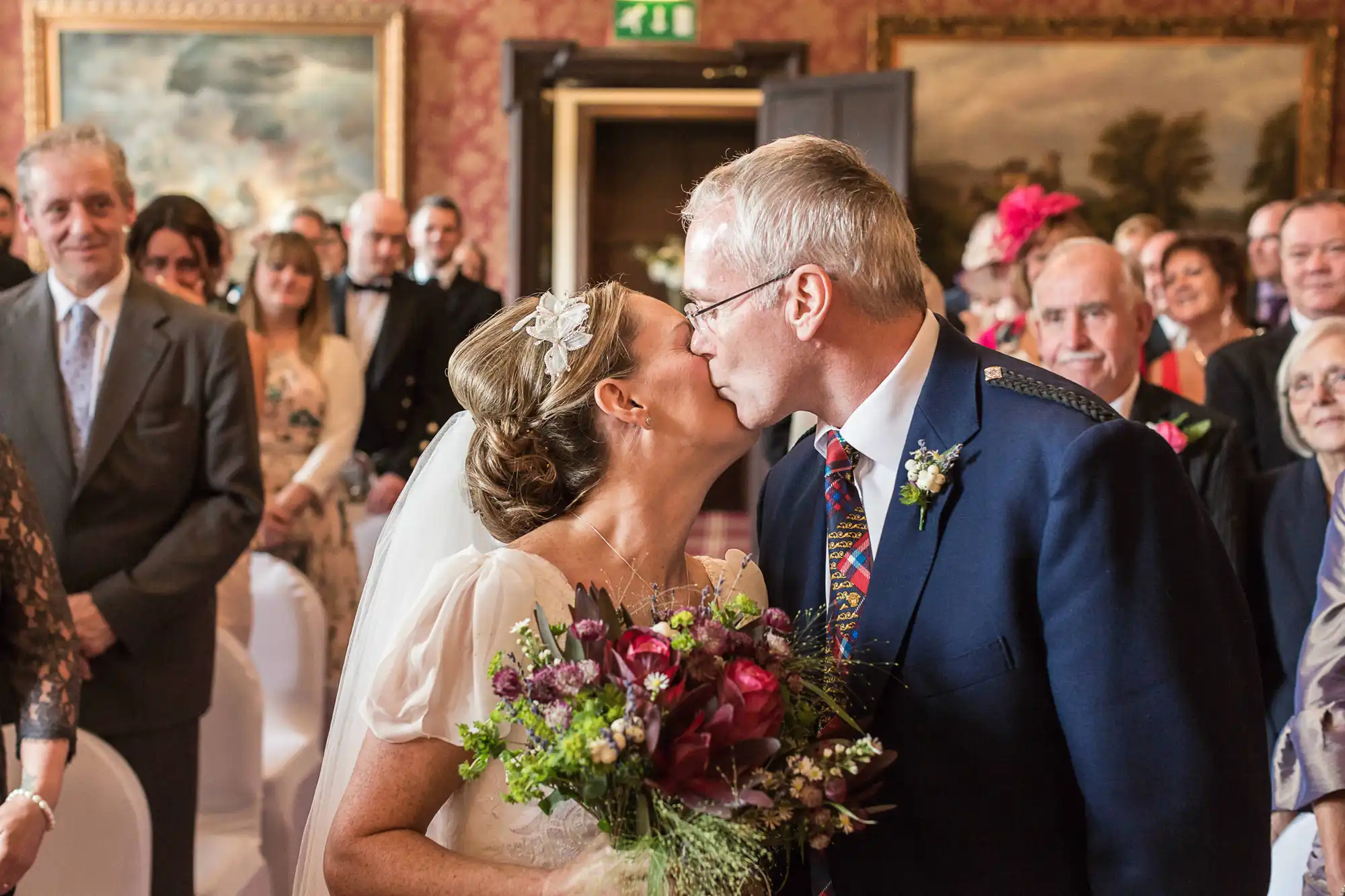 Bride and groom kissing at wedding ceremony, surrounded by guests in a decorated hall.