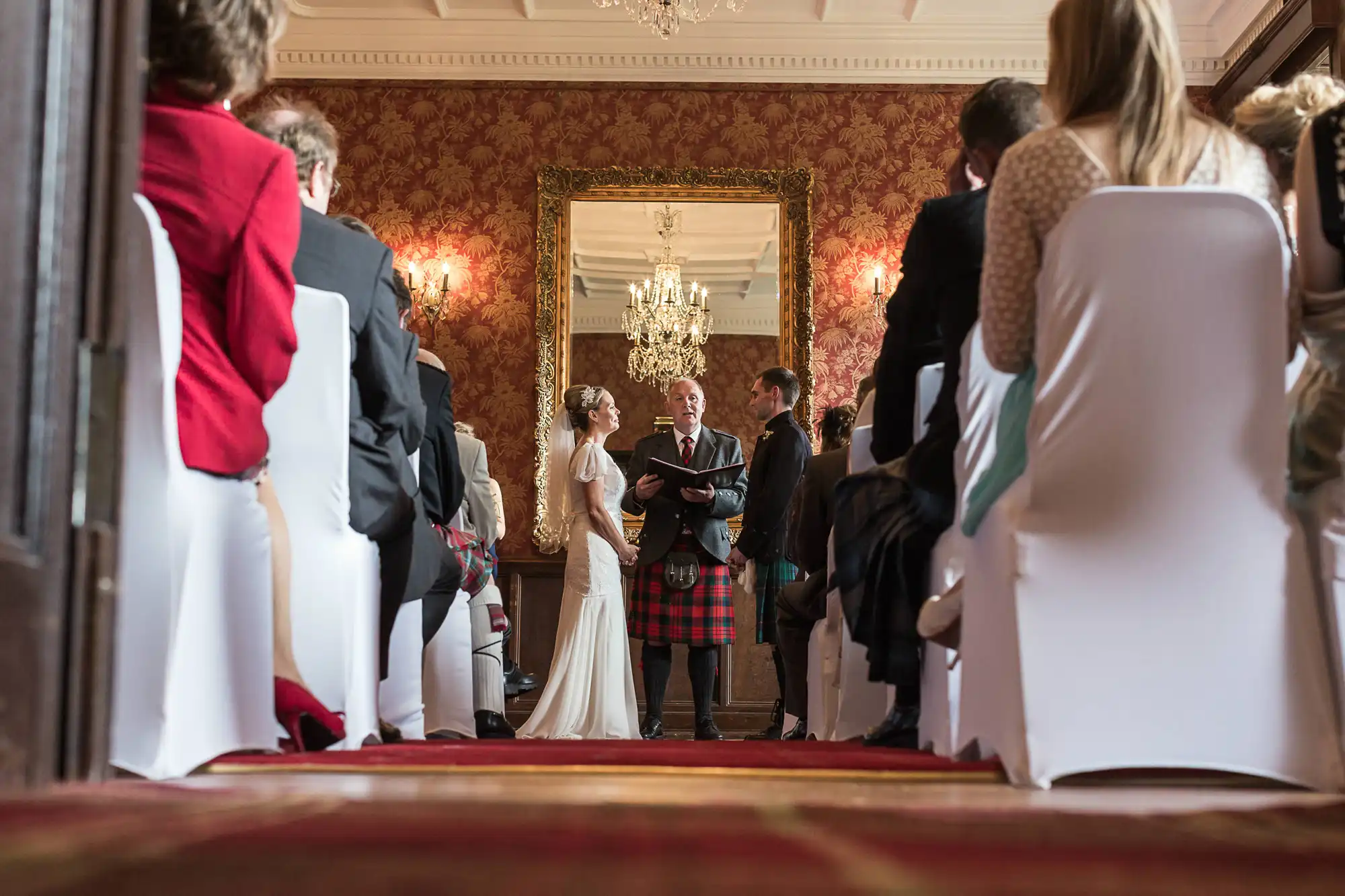 A bride and groom exchanging vows at a wedding ceremony in a luxurious room with ornate decor, observed by guests seated and standing around them.