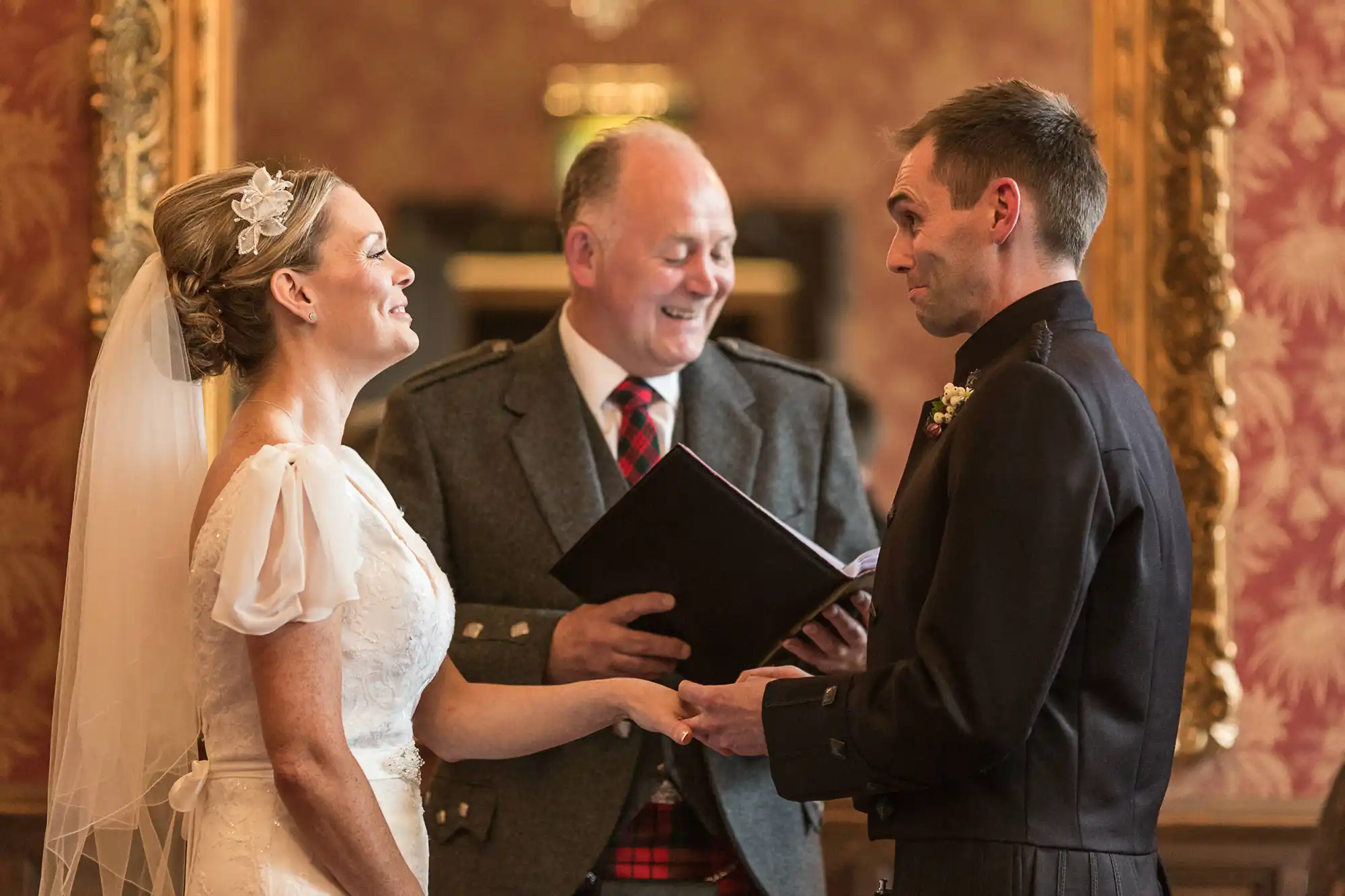 Bride and groom holding hands and smiling at each other during their wedding ceremony, with an officiant holding a book in the background.
