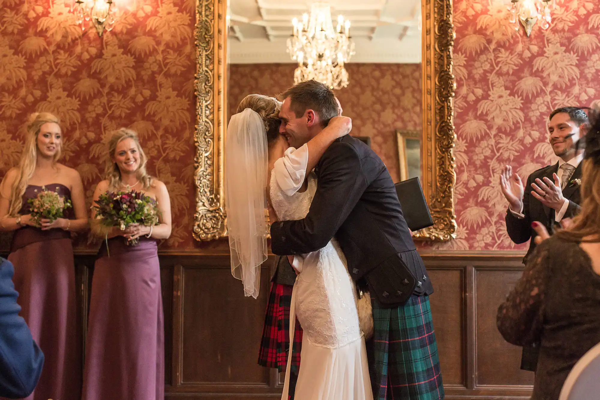 A newlywed couple kisses, surrounded by applauding guests in a vintage-style room, while a bridesmaid in purple watches.
