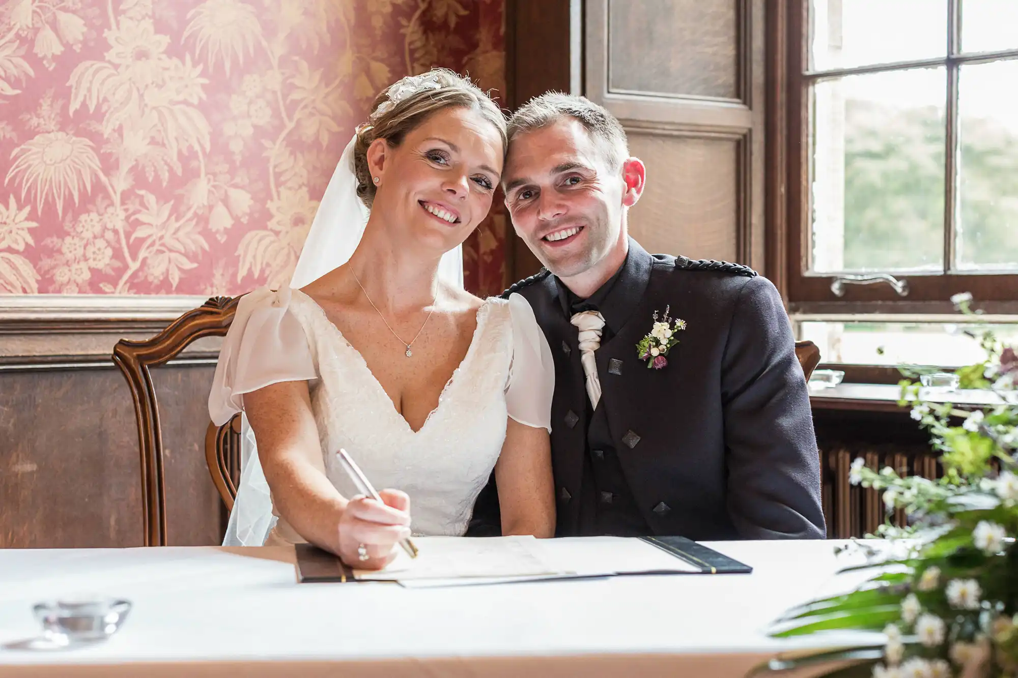 A newlywed couple signing their marriage certificate in a room with vintage decor and natural lighting.