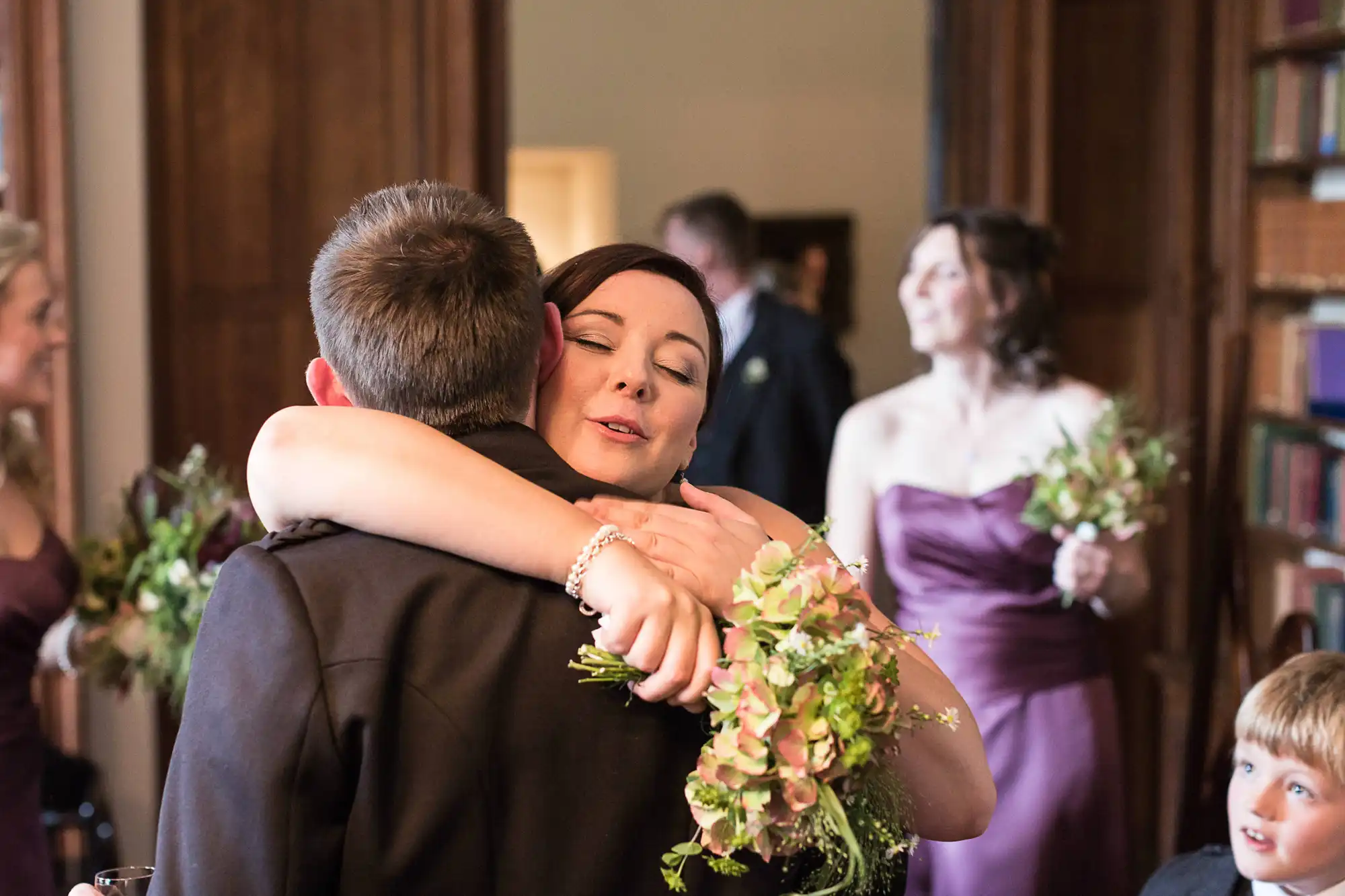 A woman in a mauve dress with a floral bouquet embraces a man in a suit at a wedding reception, while other guests look on.