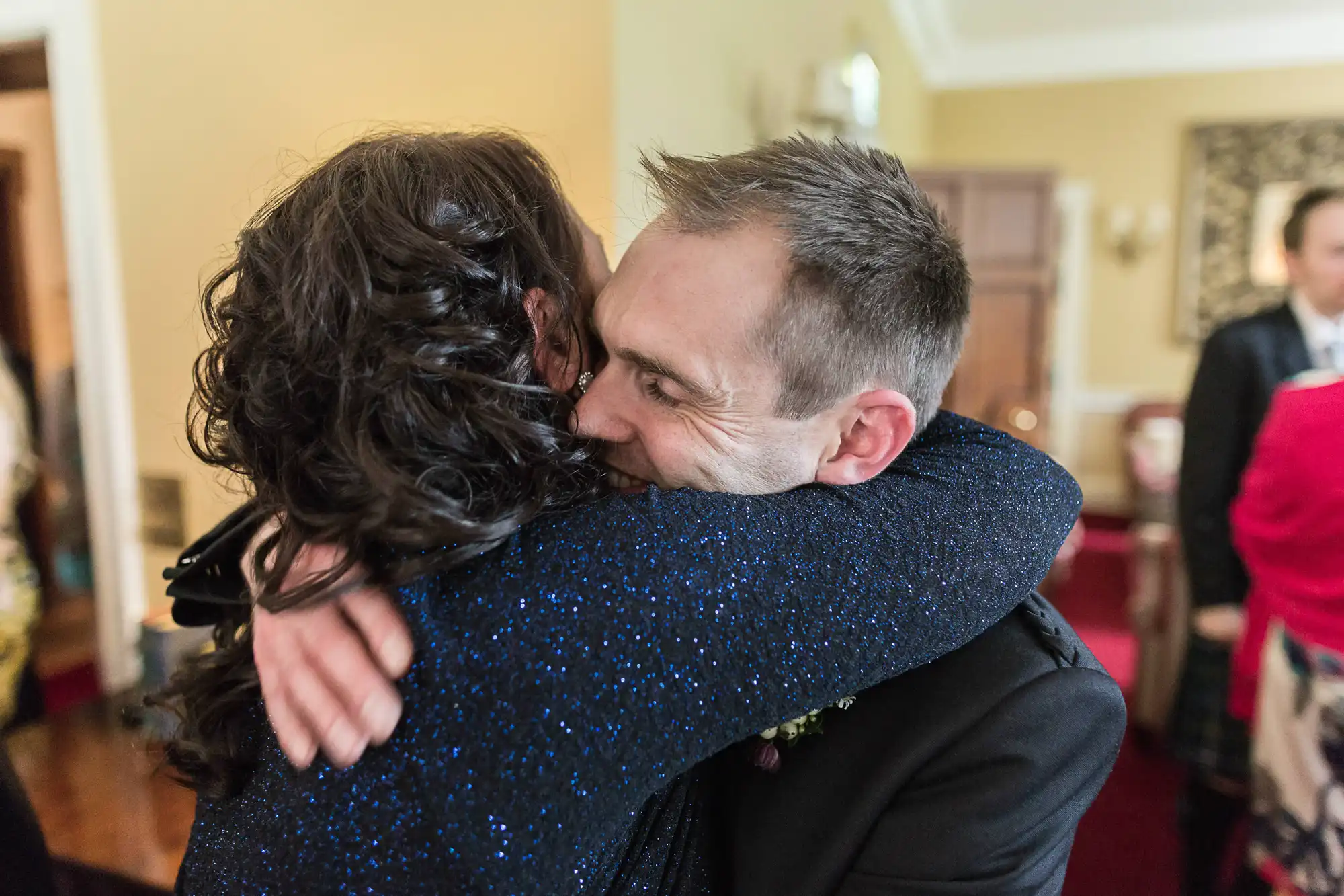 A man joyfully embracing a woman in a glittery blue dress at an indoor gathering.