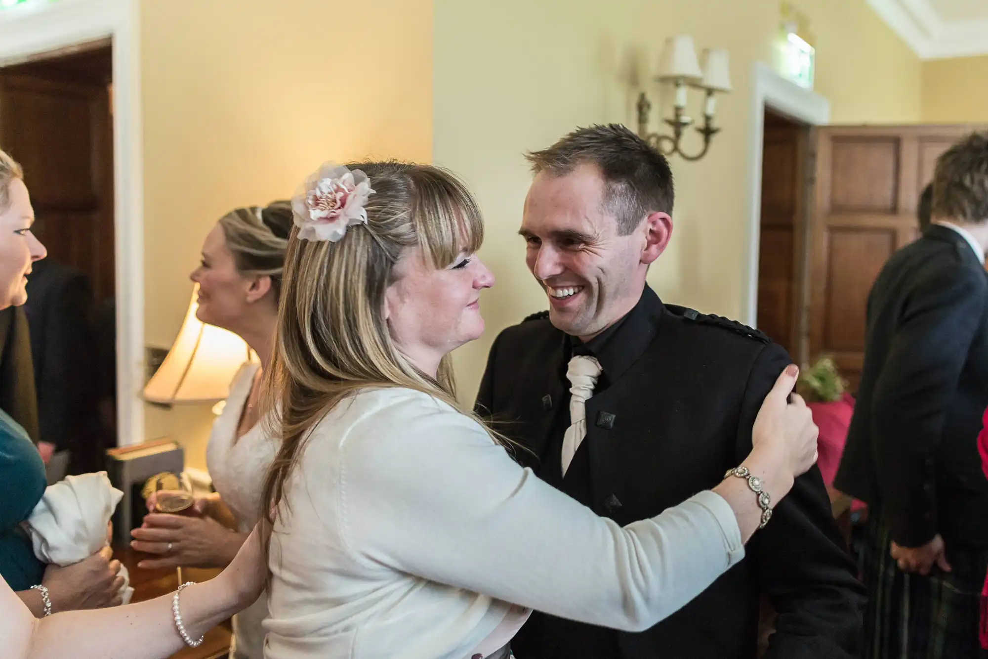 A man in a black suit and a woman with a floral hair accessory, both smiling, share a close, joyful moment at an indoor gathering.