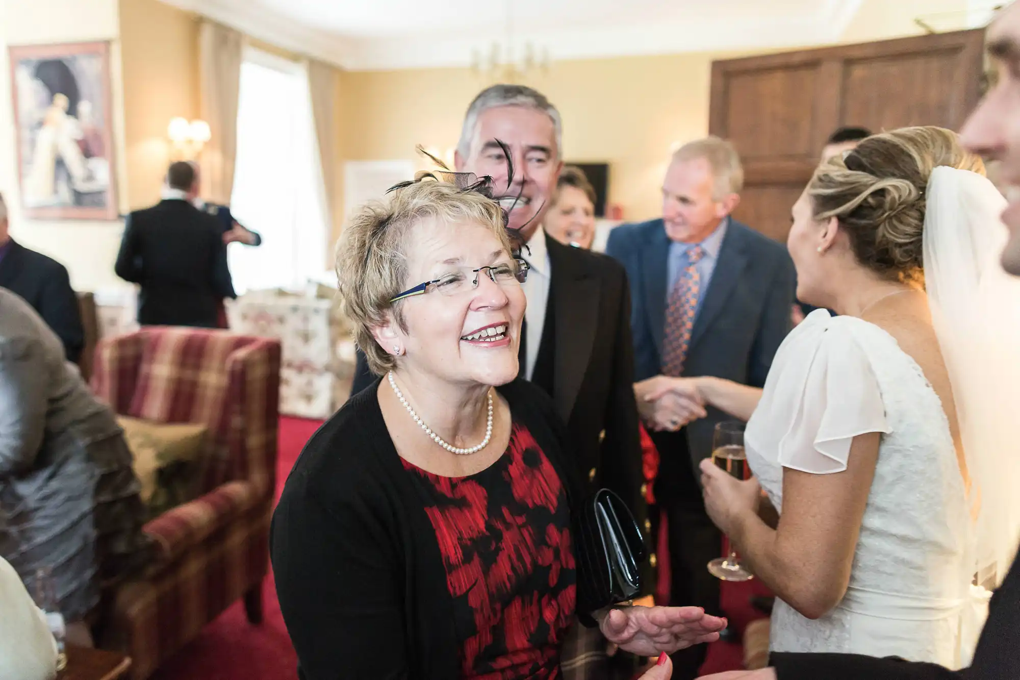 A joyful woman in a red floral dress laughs while holding glasses at a social gathering, surrounded by other guests conversing in an elegant room.