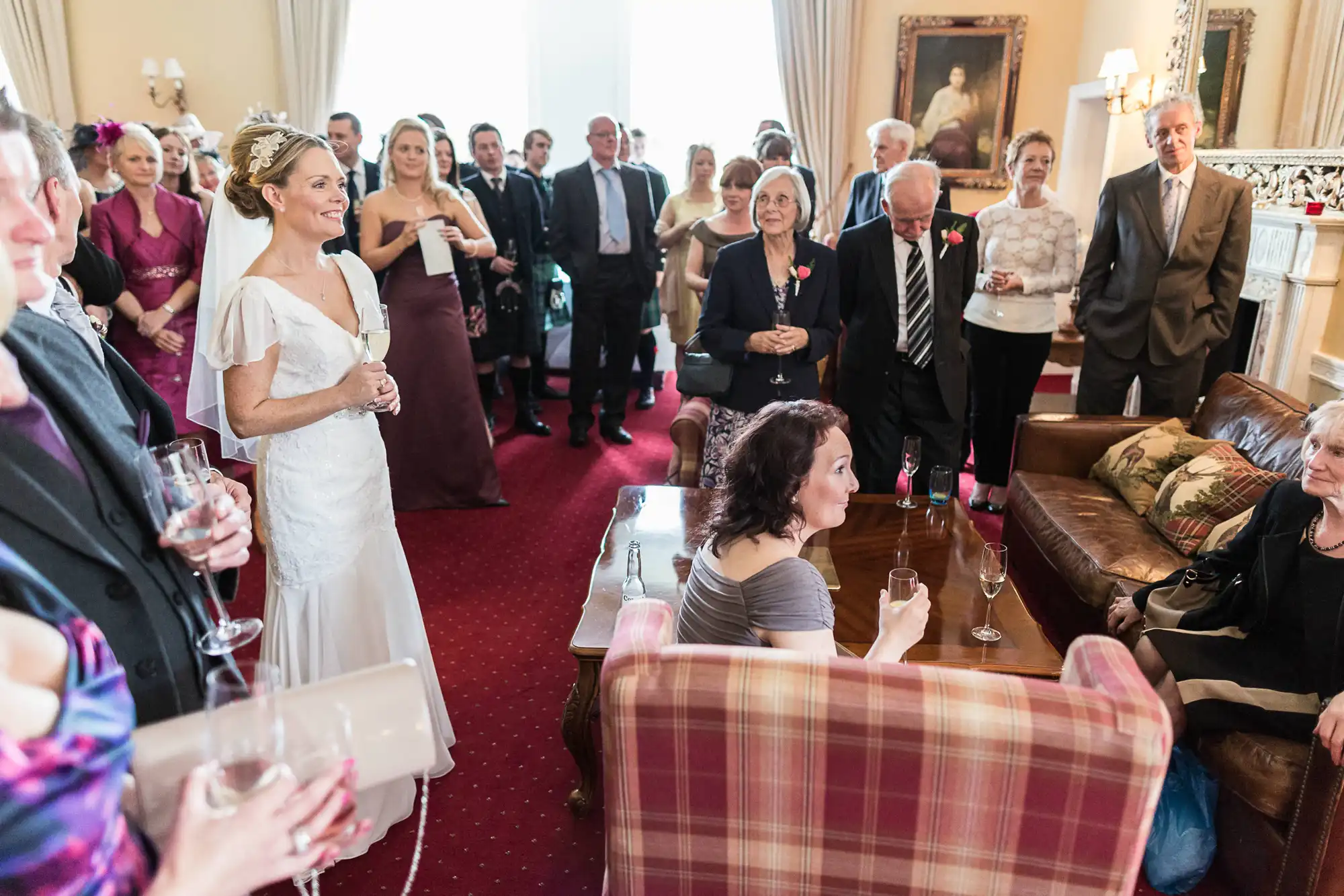 A bride in a white dress stands among guests at an indoor wedding reception, with guests in formal attire looking on and holding drinks.