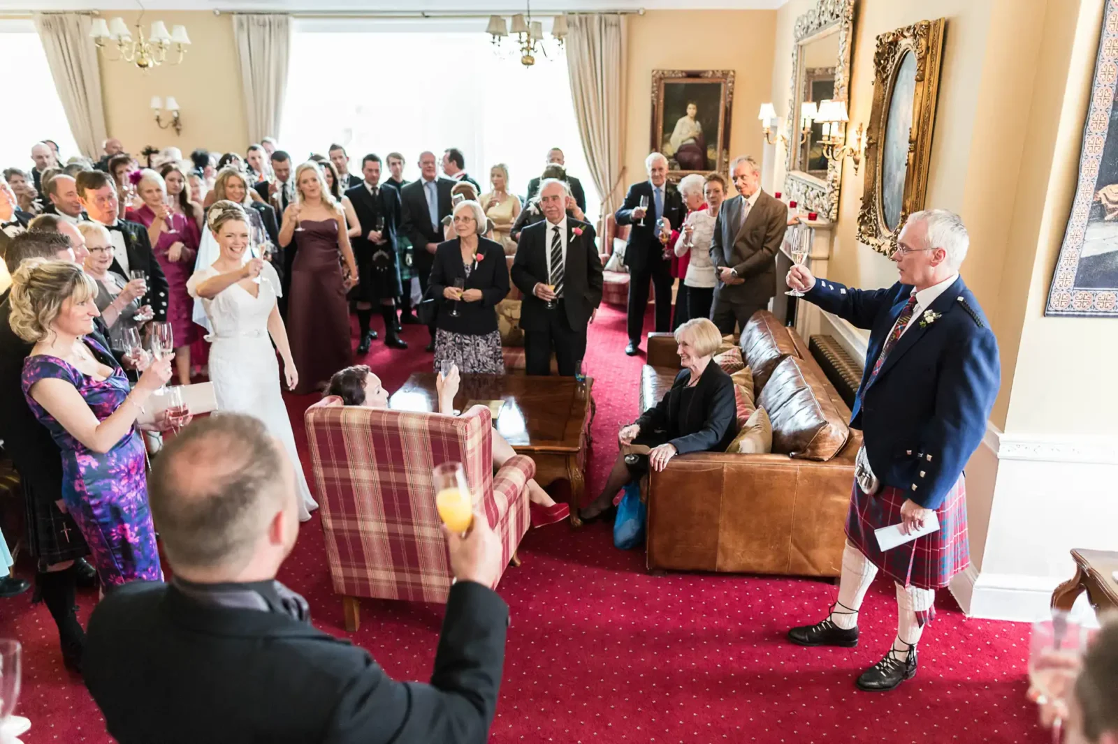 A wedding reception in an elegant room, The Drawing Room at Carberry Tower, with guests watching a man in a kilt giving a speech.