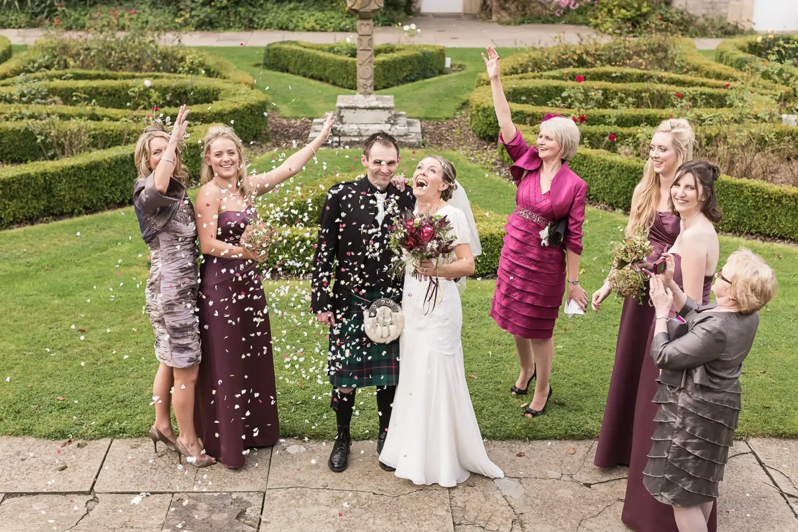 A wedding party joyfully throwing flower petals at a newlywed couple in a garden, with guests smiling and celebrating in The Rose Garden at Carberry Tower.