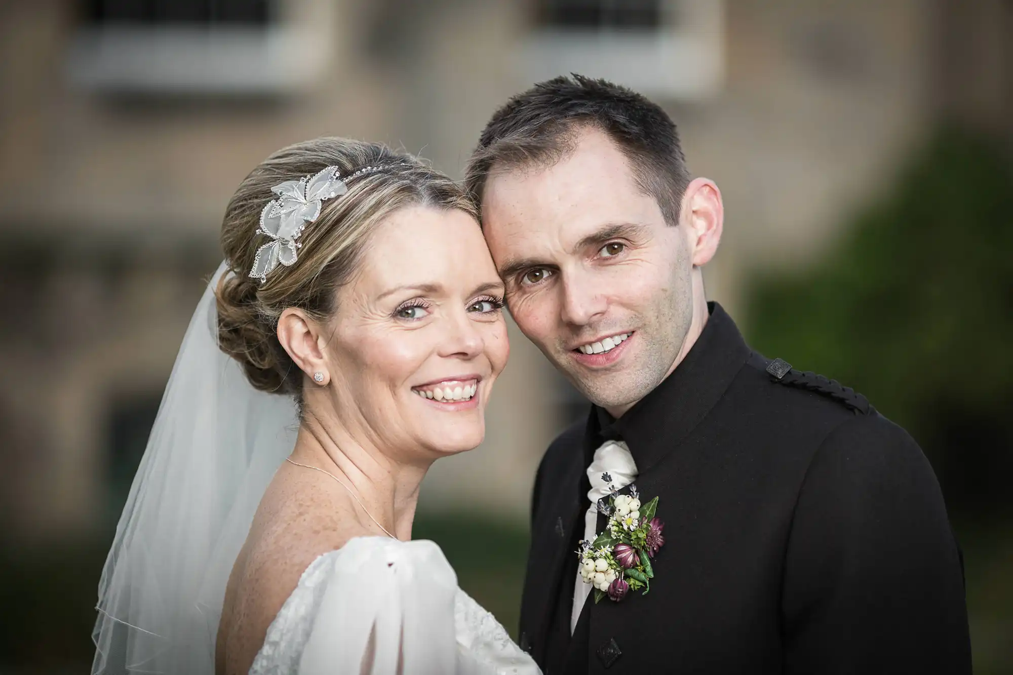 A bride and groom smiling closely together in formal wedding attire, with a historic building blurred in the background.