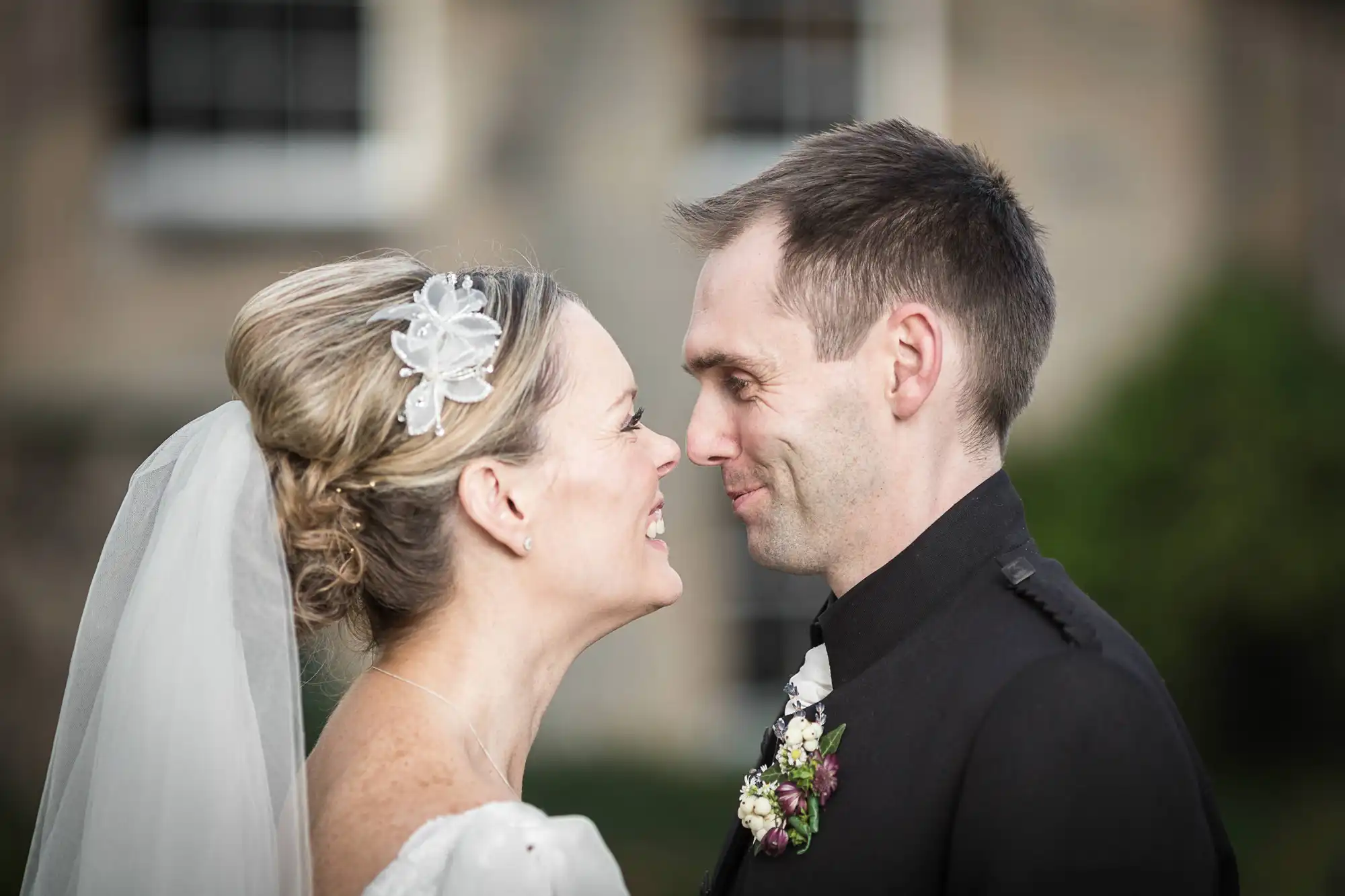 A bride and groom smiling at each other, wearing wedding attire, with a blurred building in the background.