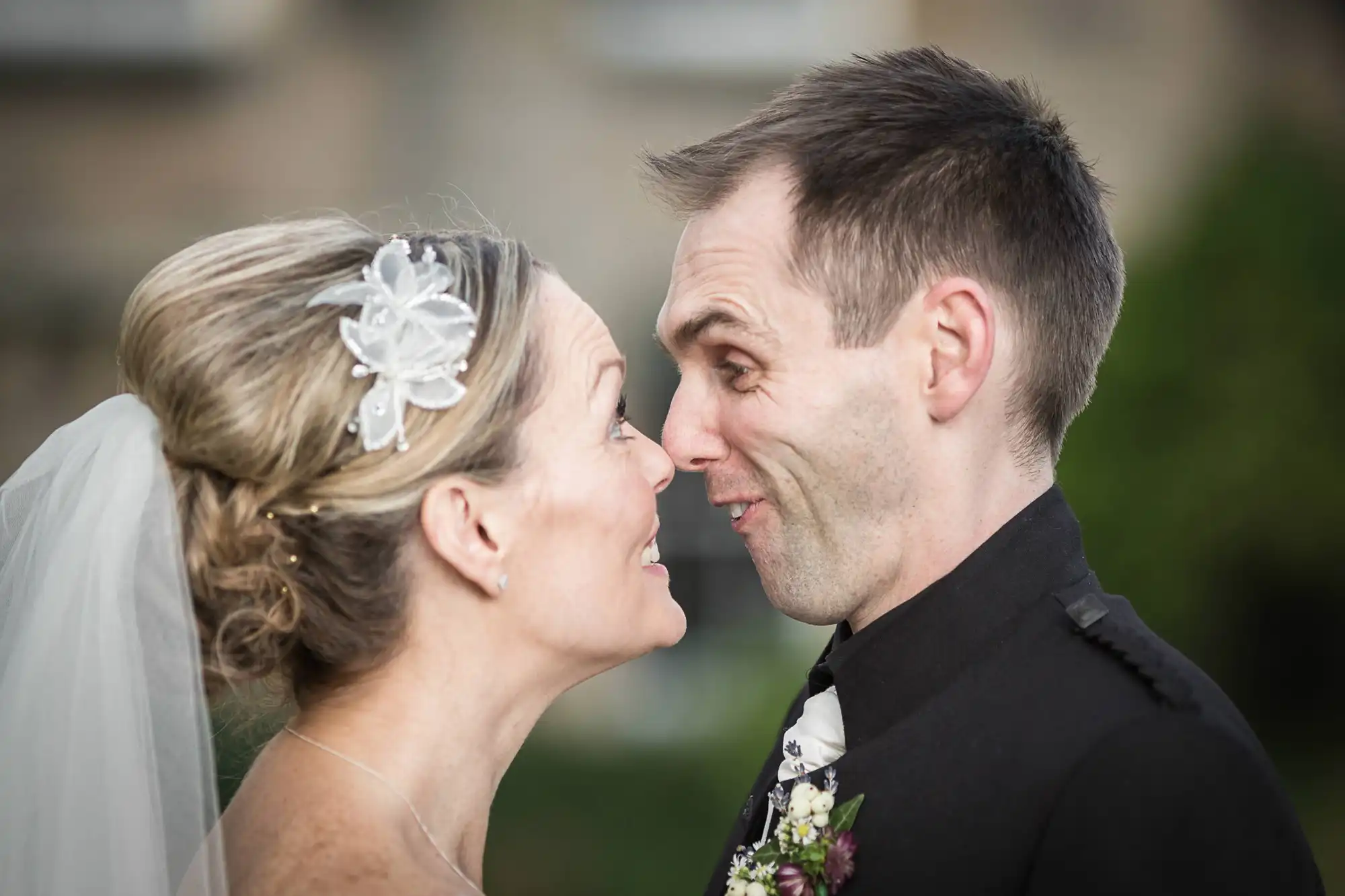 A bride and groom lovingly touch noses and smile at each other, dressed in wedding attire, with a soft-focus background in the grounds at Carberry Tower