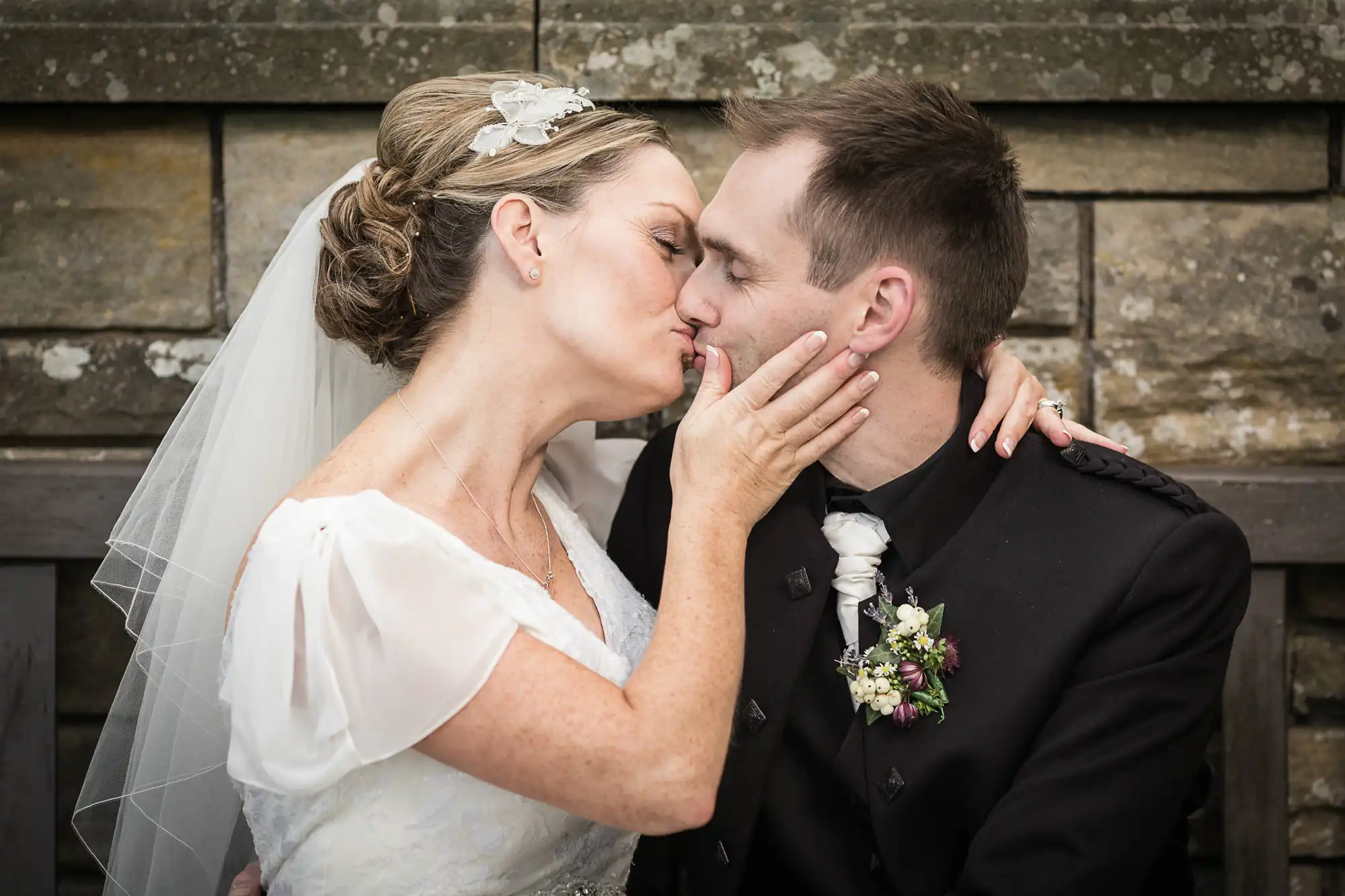 Bride and groom kissing, the bride holding the groom's face, both dressed in traditional wedding attire with a stone wall backdrop.
