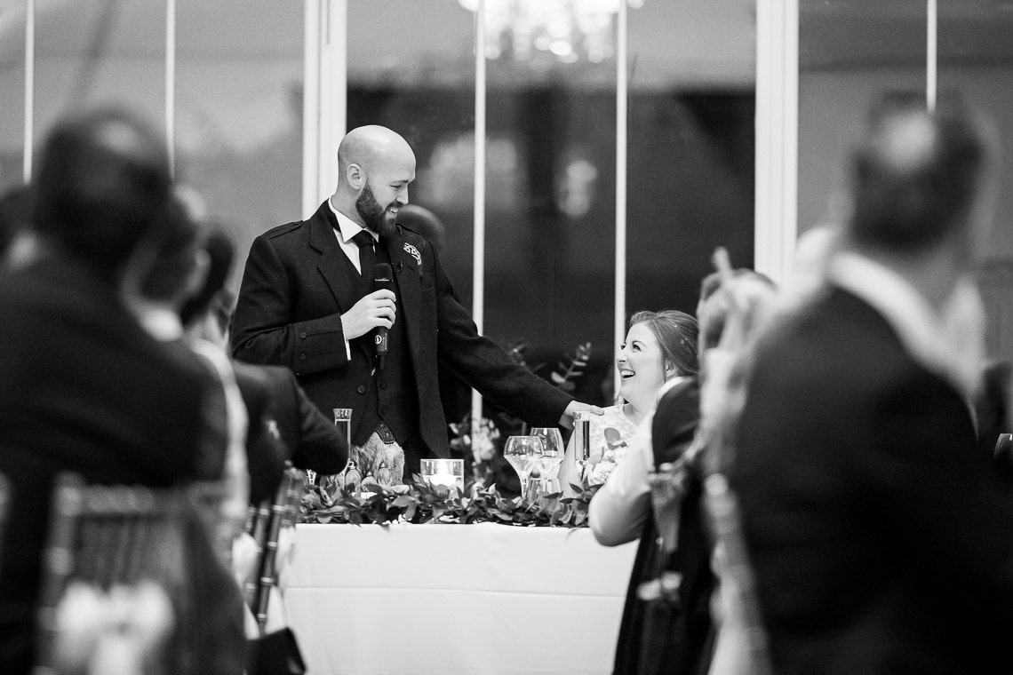 A man giving a speech at a wedding reception, holding a microphone, with a smiling woman seated at the table looking up at him; guests in the foreground are clapping.