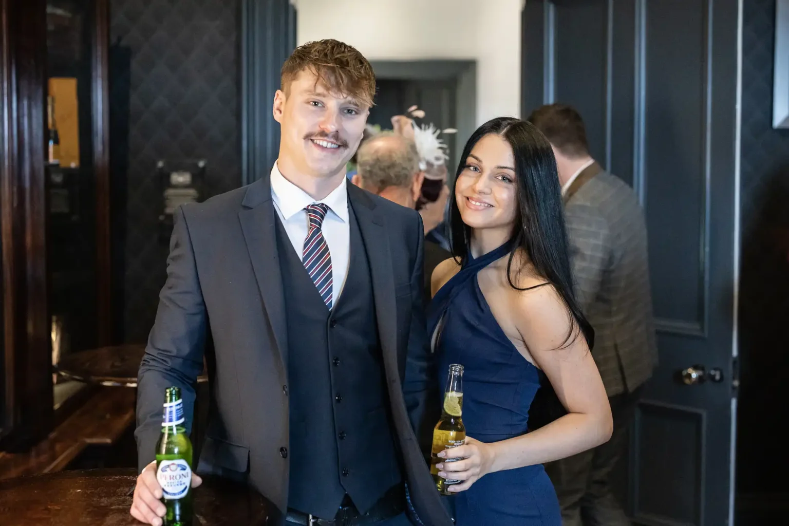 A man and a woman, dressed formally, stand side by side holding beer bottles. They are smiling and posing for the camera in an indoor setting with other people in the background.