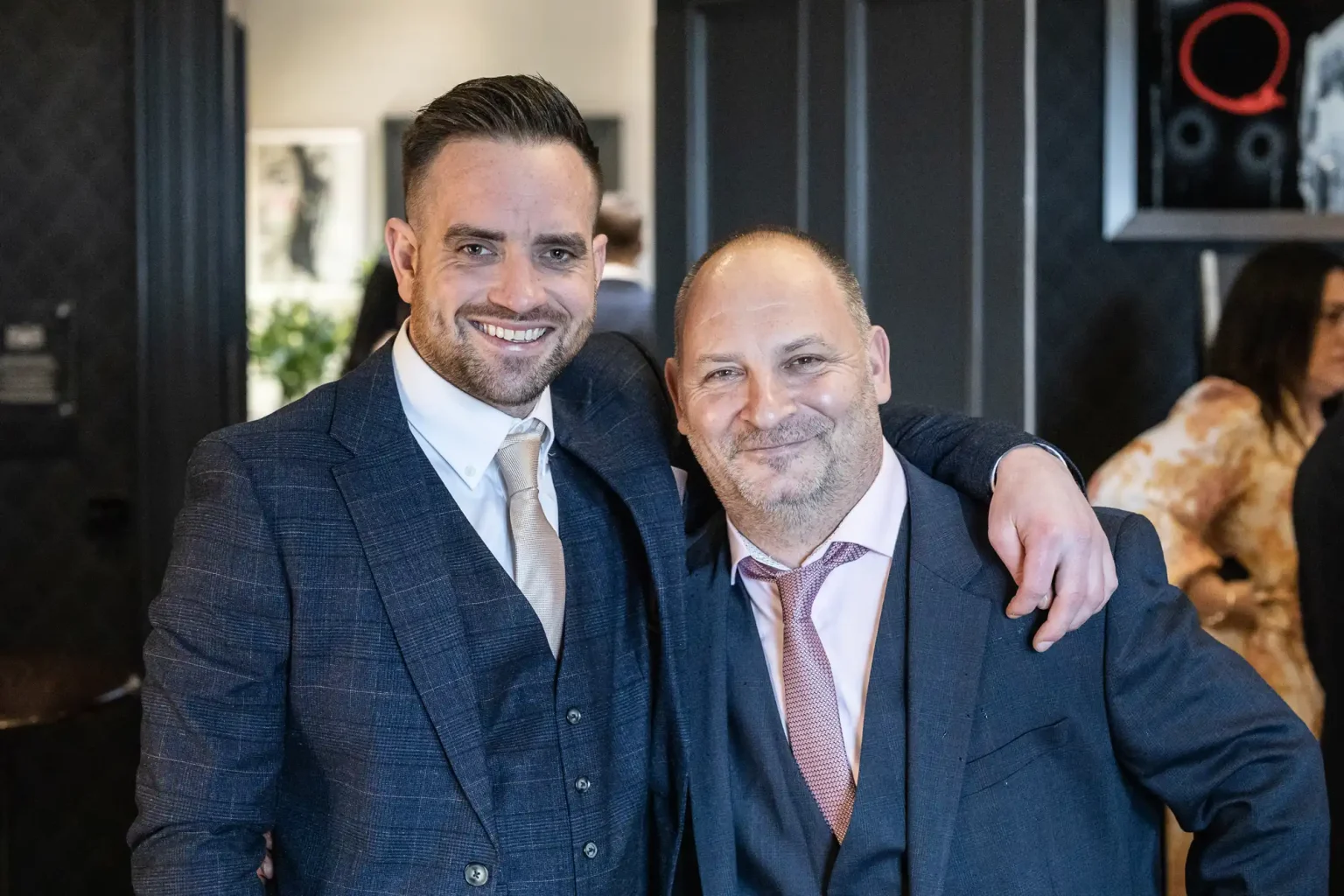 Two men in suits stand close together, smiling at the camera. One has his arm around the other. They appear to be at an indoor event or gathering, with other people visible in the background.