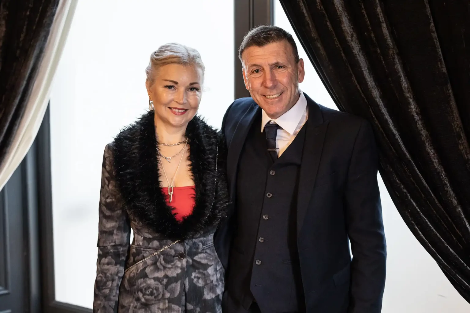 A woman in a floral dress with a black fur trim and a man in a dark suit and tie stand together near a window with dark curtains.