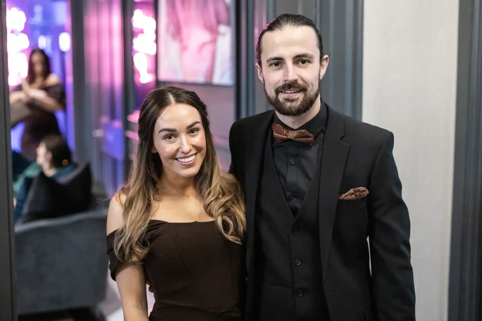 A woman and a man, both dressed in formal attire, pose together in an indoor setting. The man wears a suit and bow tie, while the woman wears an off-the-shoulder dress. Other people are in the background.