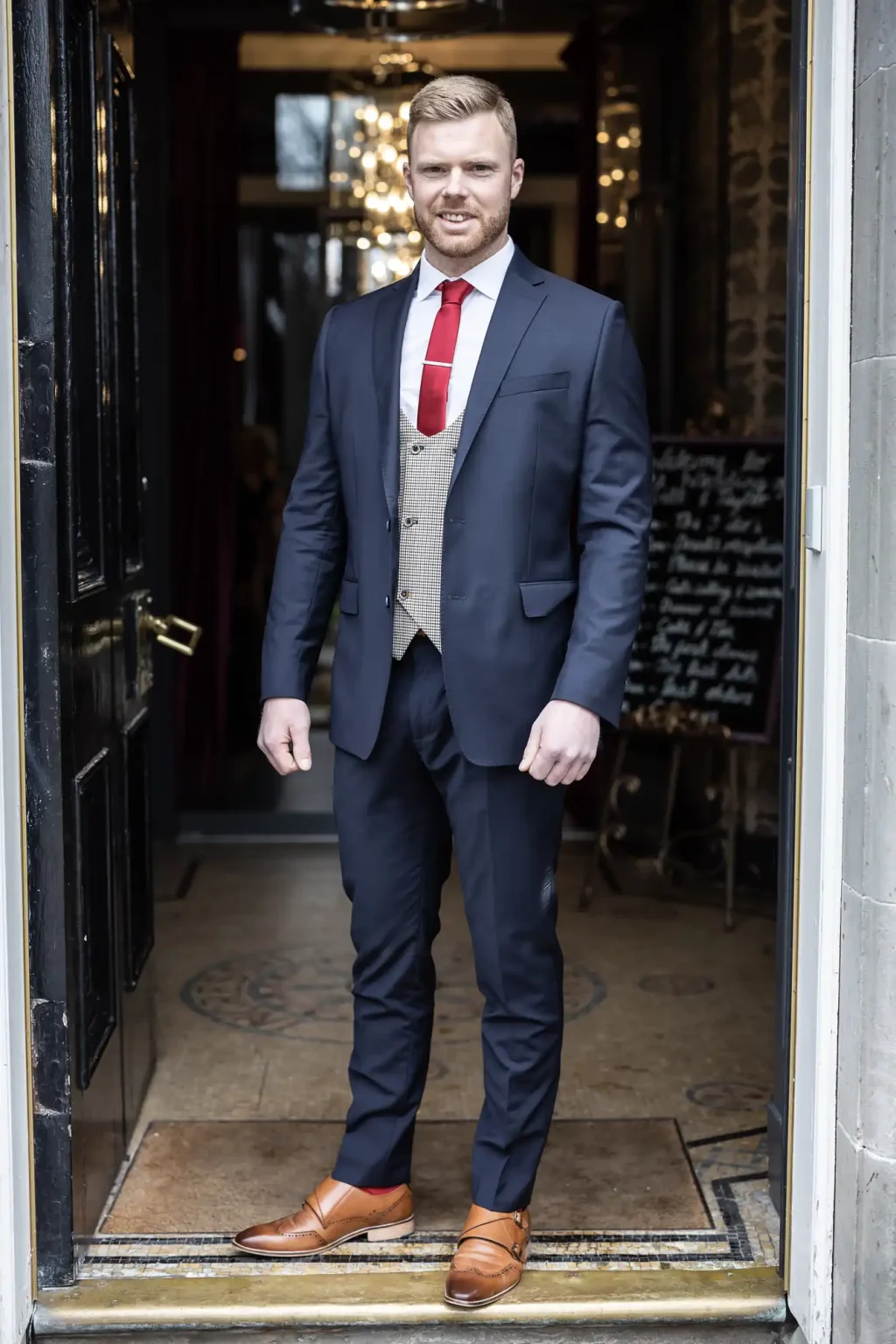 A man with short hair and a beard stands in a doorway wearing a dark suit, red tie, and light brown shoes, looking toward the camera.
