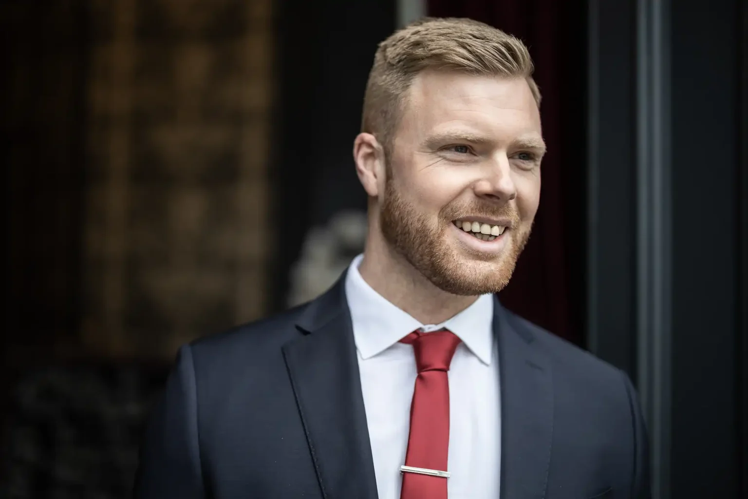 A man with short, light brown hair and a beard, wearing a dark suit, white shirt, and red tie, smiles while looking to the side.