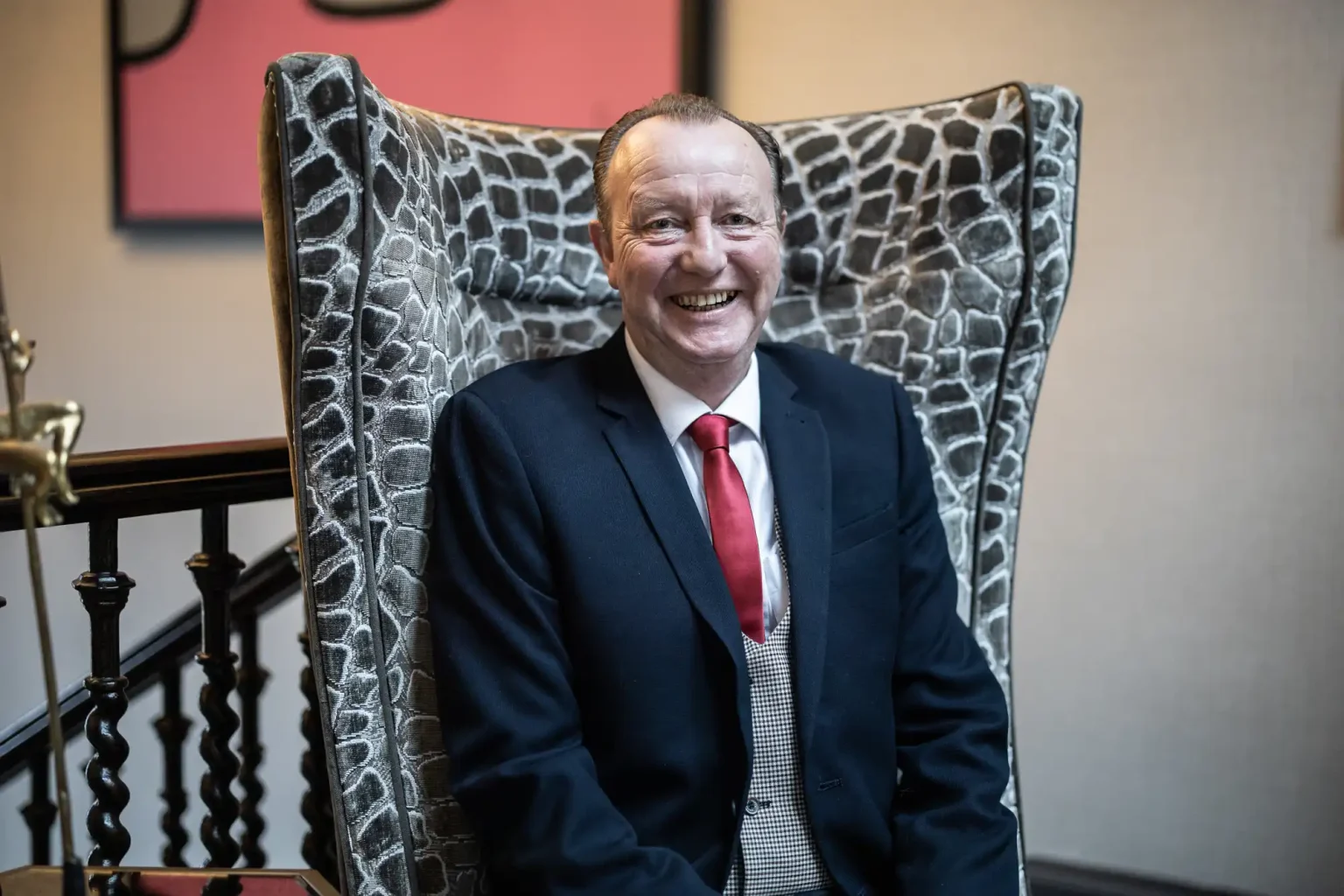 A man in a suit and red tie sits on a patterned high-back chair, smiling. He is indoors with a staircase and a pink artwork in the background.