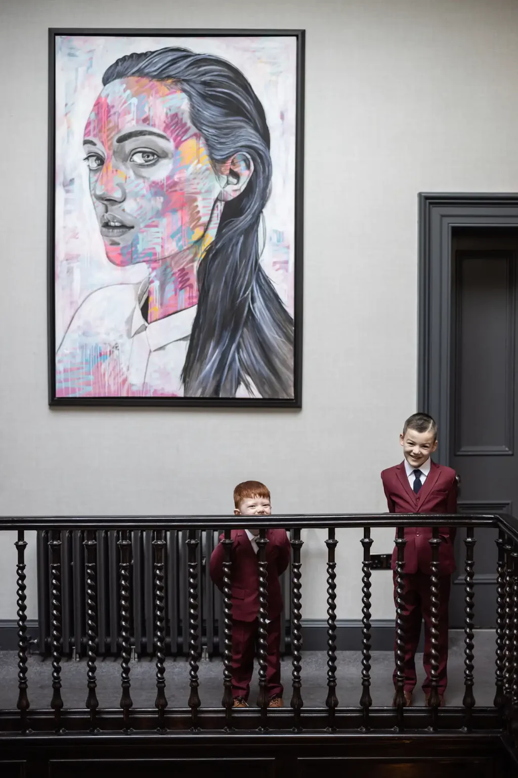 Two boys in red suits stand beside a dark wooden railing; a colorful portrait of a woman is hung on the wall behind them.