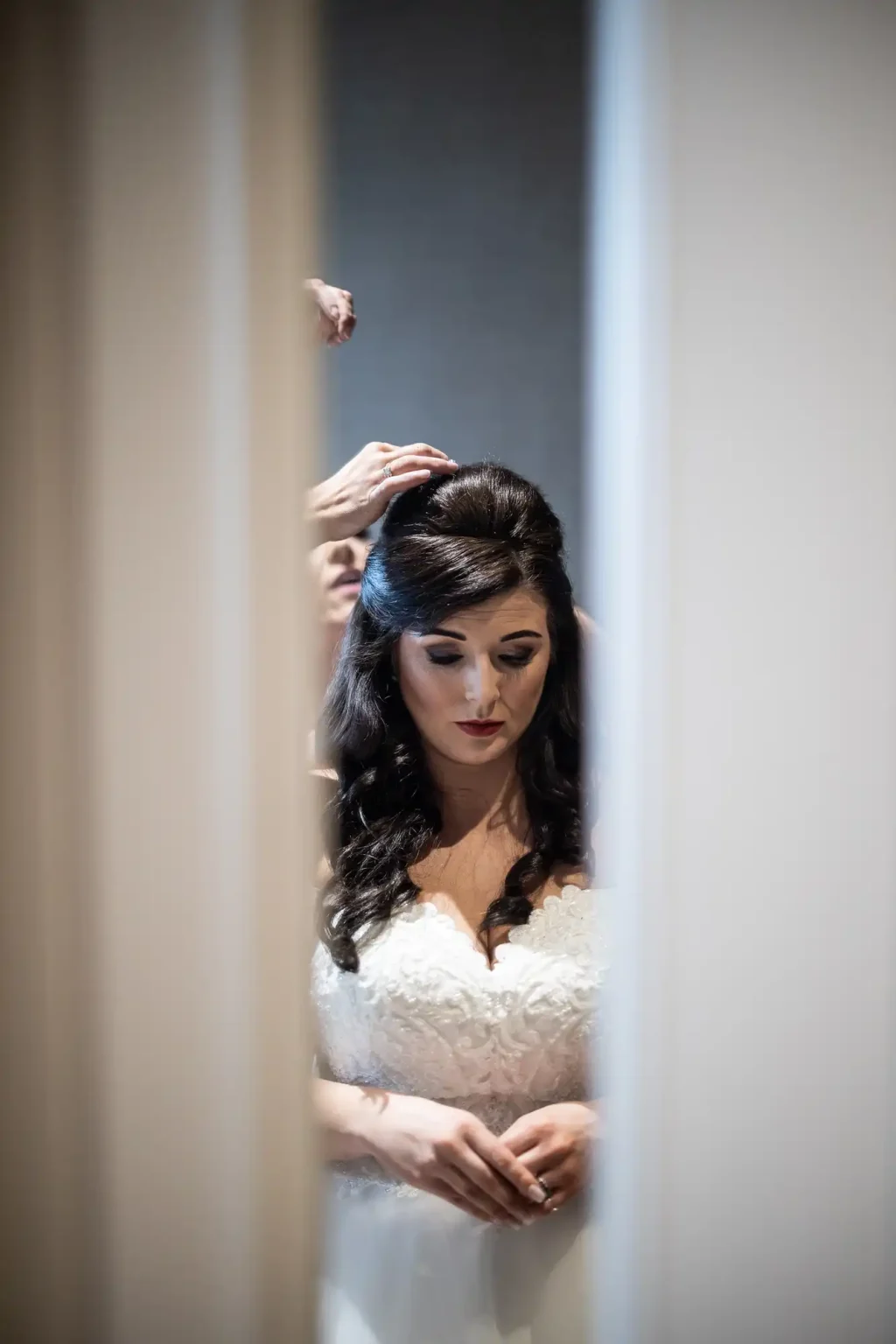 A bride in a white wedding dress stands with hands clasped, her dark hair being styled by someone behind her.