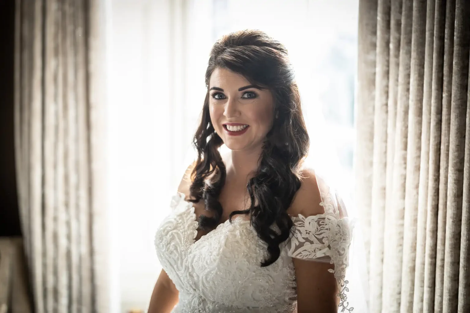 A woman in a bridal gown stands between two curtains, smiling at the camera.