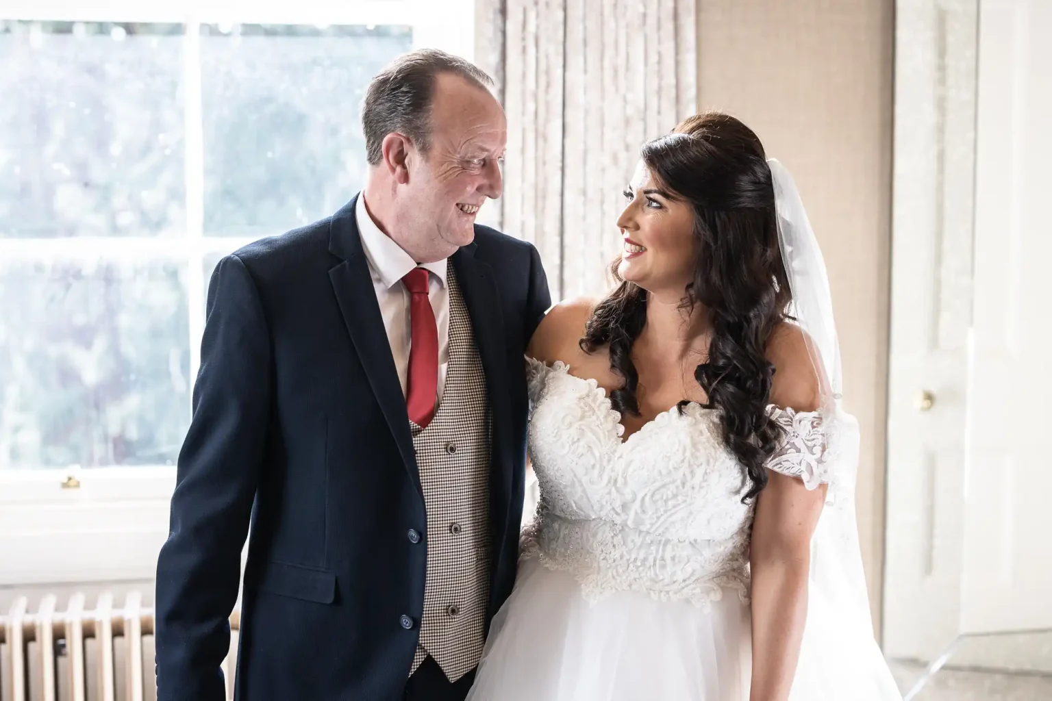 A bride in a white wedding dress and veil stands indoors, smiling at an older man in a suit and red tie. They look at each other with a window in the background.