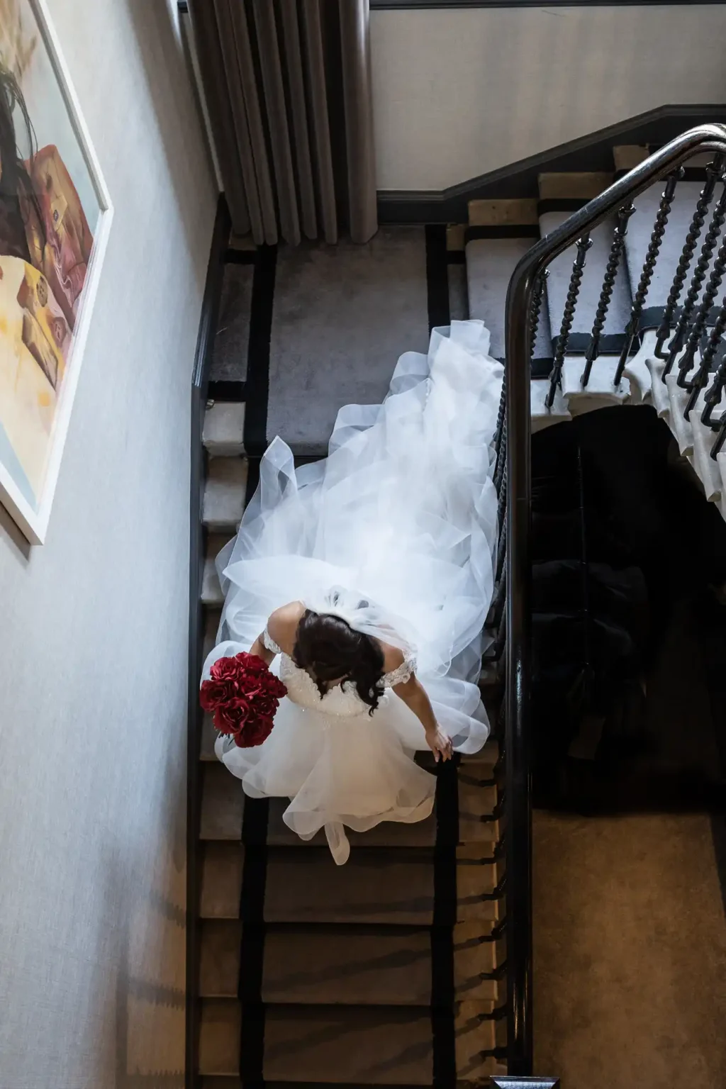 A bride in a white wedding dress with a long train descends a staircase while holding a bouquet of red flowers.