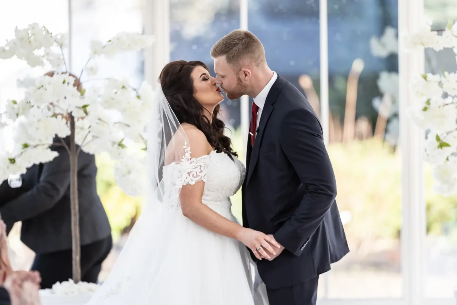 A bride and groom share a kiss during their wedding ceremony, standing between white floral arrangements.