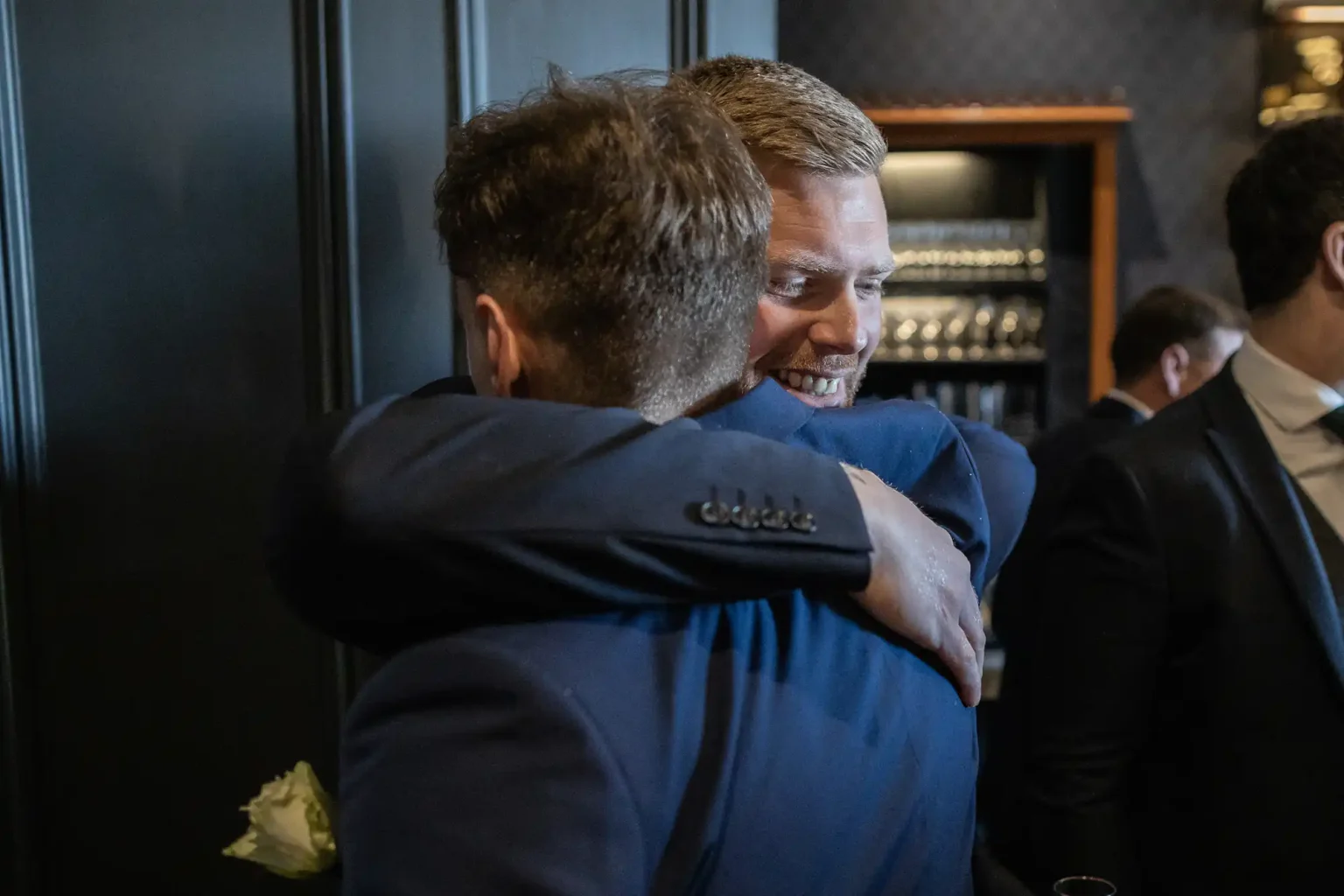Two men in suits embracing while smiling in a dimly lit room, with other people blurred in the background.