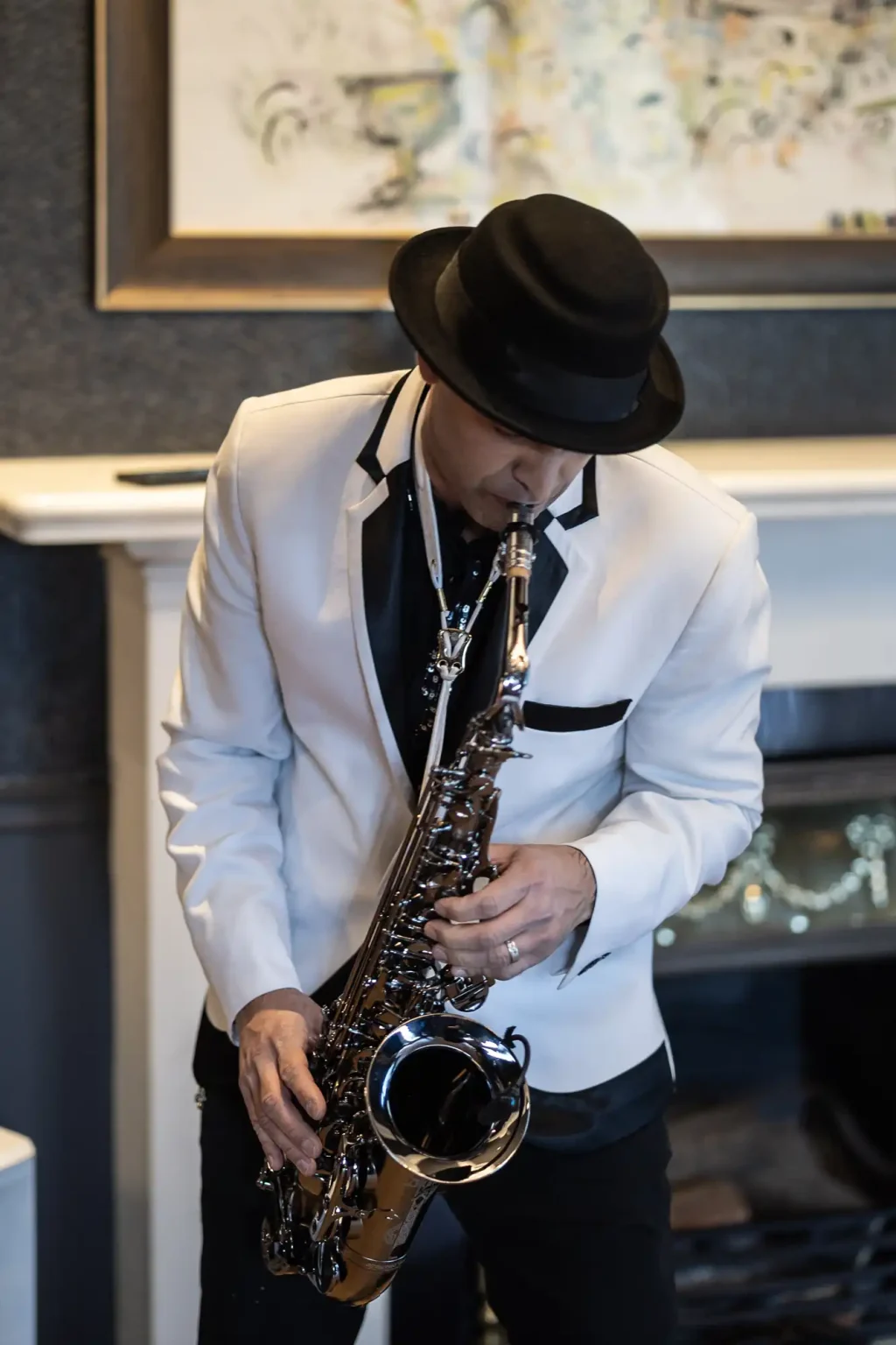 A man in a white suit jacket and black hat plays a saxophone indoors beside a fireplace.