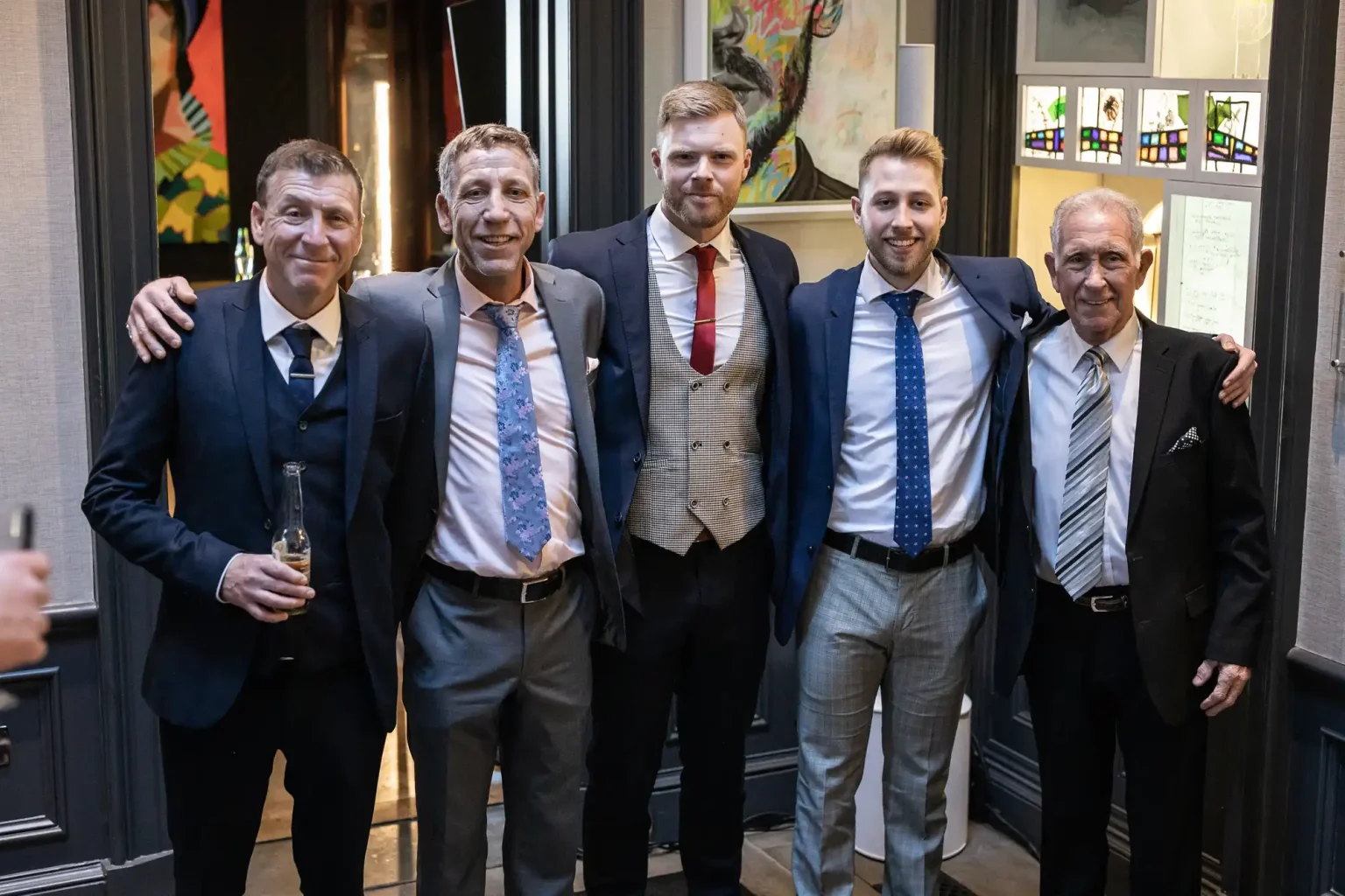 Five men in suits pose together in a formal setting, with one of them holding a drink. They are standing in a line, smiling at the camera.