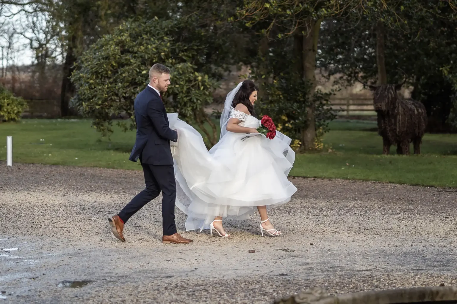 A bride in a white gown and veil holds a bouquet of red flowers while walking on a gravel path with a groom wearing a dark suit. Trees and a metal sculpture of an animal are in the background.