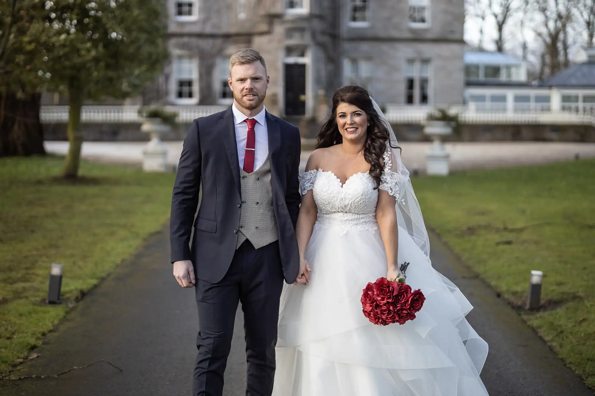 A couple dressed in formal attire stands outdoors holding hands. The man wears a dark suit with a red tie, and the woman wears a white wedding dress and holds a red bouquet. A building is in the background.