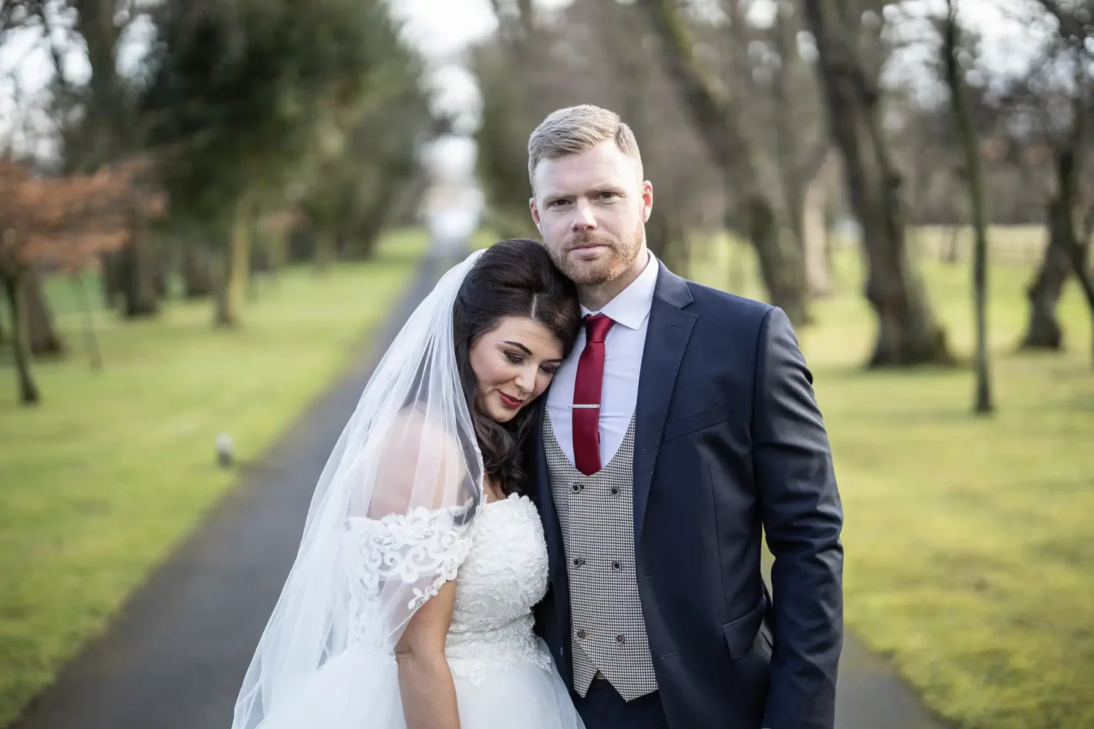A bride in a white dress and veil leans her head on the shoulder of a groom in a dark suit and red tie, standing on a tree-lined path.