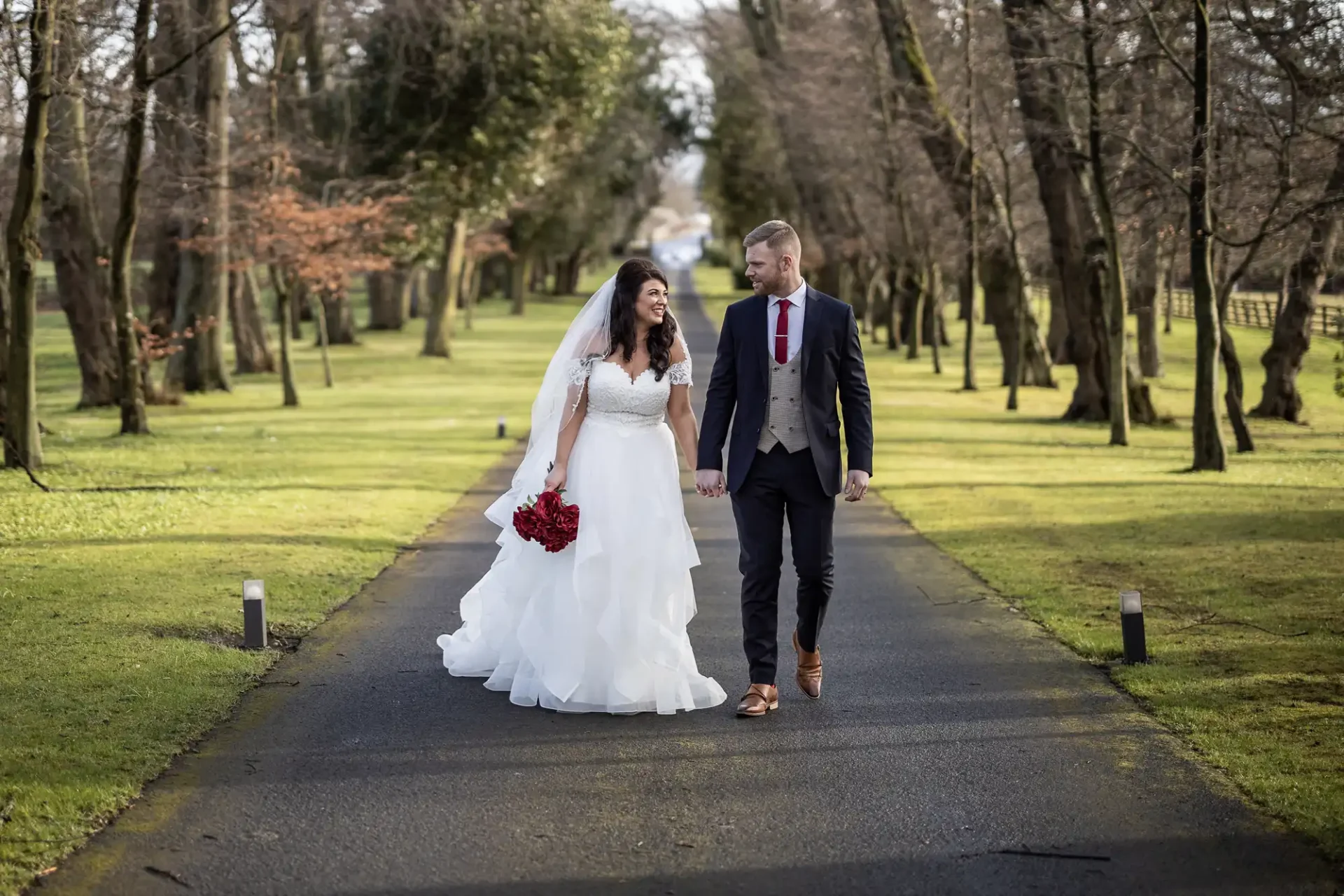 A couple in wedding attire walks hand in hand down a tree-lined drive at Carlowrie Castle. The bride holds a red bouquet and wears a white dress with a veil, while the groom wears a suit with a red tie, featured on a page about Carlowrie Castle weddings.
