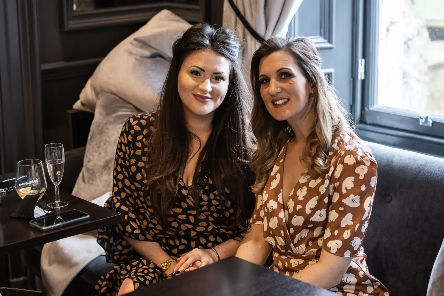 Two women sitting together at a table, each with a glass of champagne in front of them. They are both smiling and wearing patterned dresses.