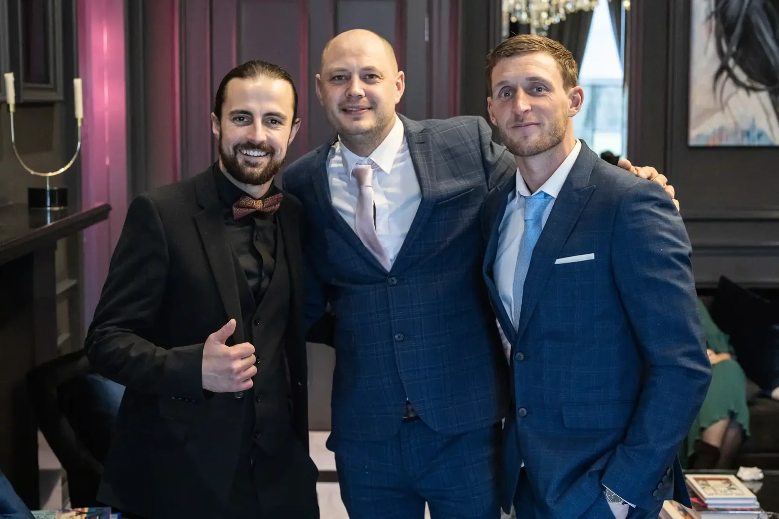Three men are dressed in suits, posing together indoors. The man on the left is giving a thumbs up, the man in the middle has his arm around the other two, and the man on the right is standing close.