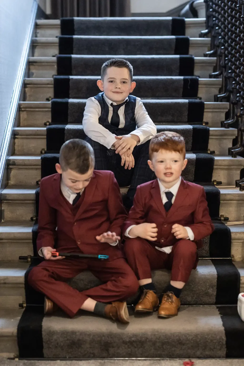 Three boys sit on a staircase. The boy at the top is in a white shirt and black vest, while the two boys below are wearing maroon suits. One of the boys in a maroon suit is using a tablet.