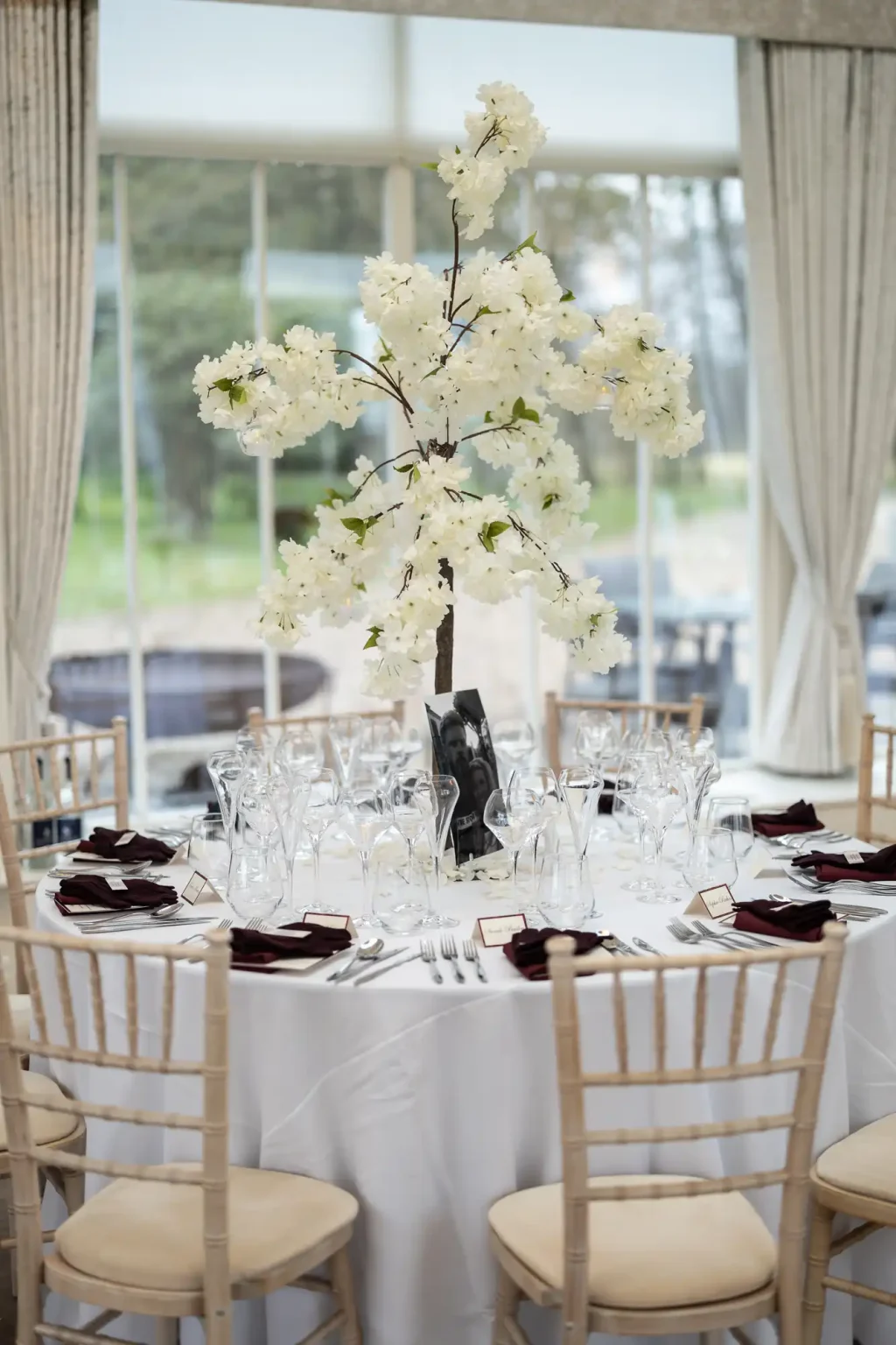 A round table with a white tablecloth, set for an event, featuring a tall floral centerpiece with white flowers, surrounded by wine glasses and burgundy napkins.