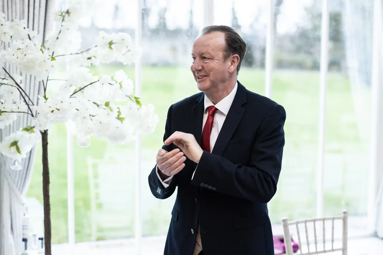 An older man in a black suit with a red tie stands indoors near a white-flowered plant, smiling and holding an object in his right hand.